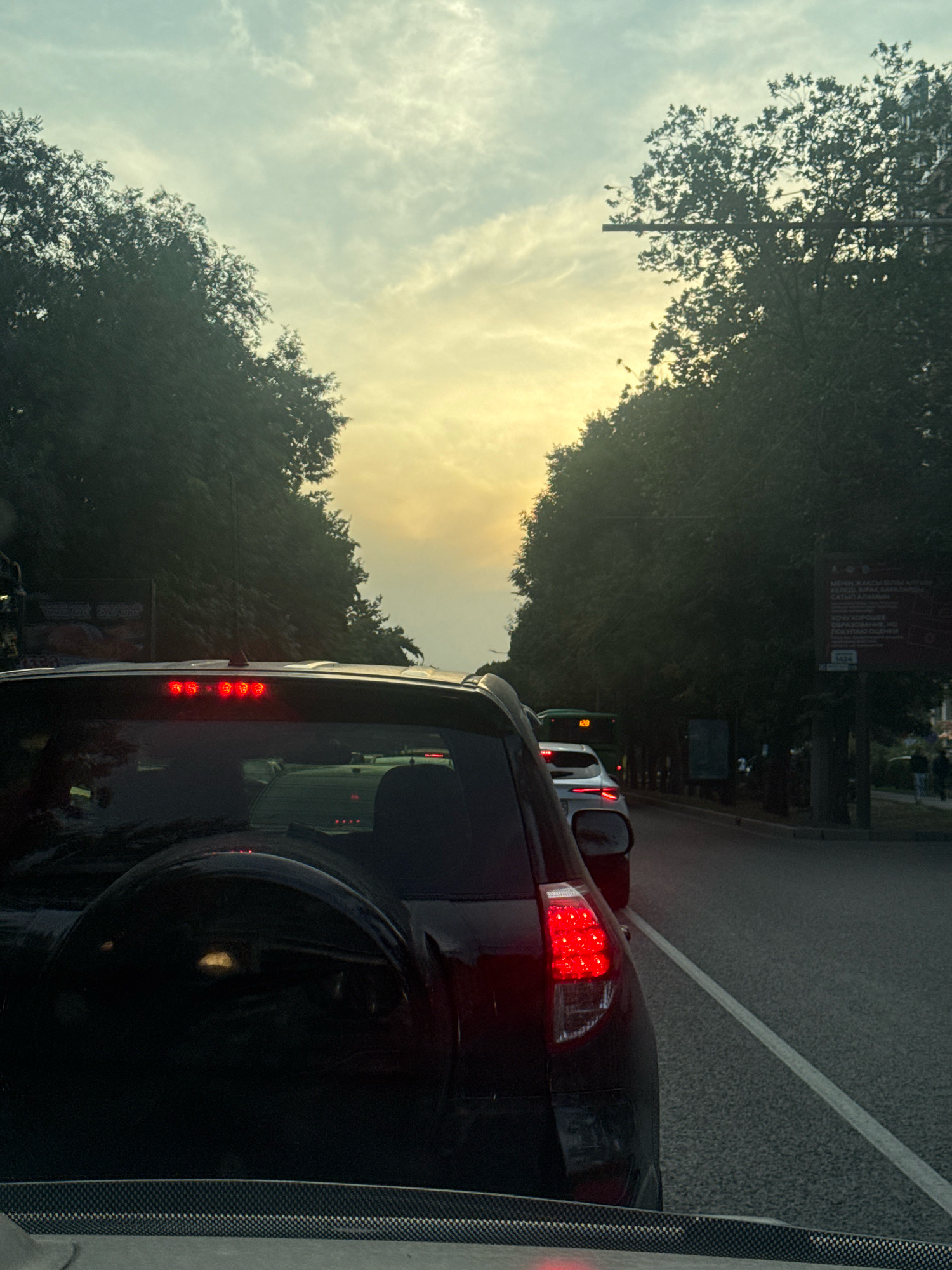 The image captures a street scene in Almaty, Kazakhstan, characterized by heavy traffic and a cloudy sky tinged with yellow light, suggesting either dawn or dusk. The perspective is from within a vehicle, looking forward through its rear window and out at the road.
In the foreground, the rear of a dark-colored SUV is prominent, with its brake lights illuminated red, indicating it is stopped or slowing down. A spare tire is mounted on the back of this vehicle. The vehicle in front of it is a white SUV, also stopped in traffic. Further ahead, a green bus is visible, displaying the number "128" on its front.
The street is lined with dense, dark green trees on both sides, creating a canopy effect over the road. There is a sidewalk on the right side of the street, where a few pedestrians are walking. A large billboard with text in what appears to be Cyrillic script is mounted on a pole beside the road, partially obscured by trees. The road surface is asphalt, marked with white lines. The overall impression is one of a typical urban traffic jam in a city with significant tree cover.