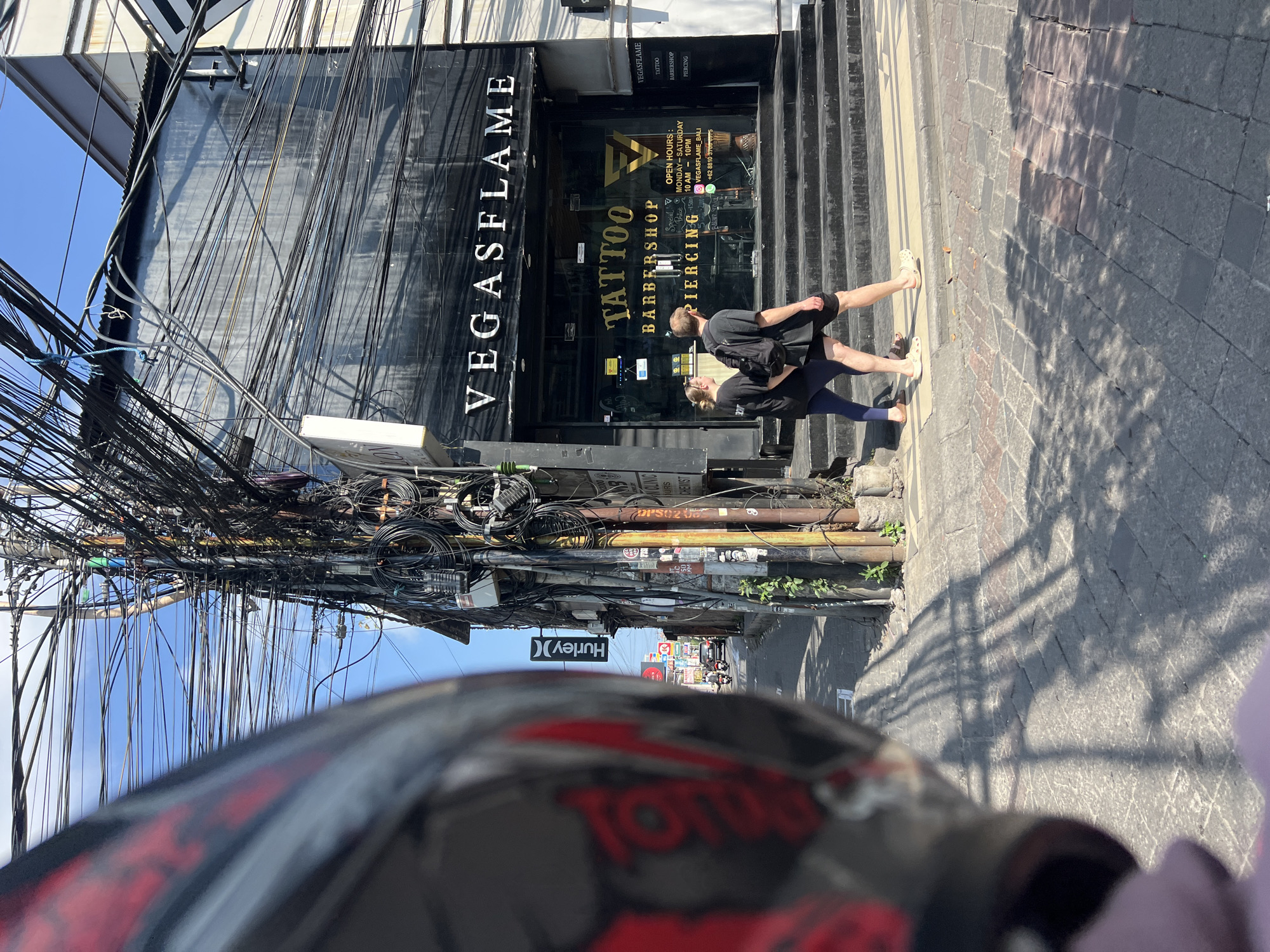 In Kuta, Indonesia, on a sunny day, a street scene unfolds with a prominent tattoo and barbershop named "VEGASFLAME" as the focal point. The building's facade features large glass windows showcasing the shop's interior and a sign that reads "TATTOO BARBERSHOP PIERCING." Above the entrance, the name "VEGASFLAME" is displayed in large white lettering. Two individuals, a man and a woman, are seen walking across a pedestrian crossing in front of the establishment. The man, wearing a black t-shirt and shorts, carries a backpack. The woman, in a black t-shirt and blue leggings, walks beside him. The street is paved with cobblestones, and overhead, a dense network of tangled electrical wires creates a chaotic canopy against the bright blue sky. The foreground of the image is partially obscured by the blurred reflection of a motorcycle helmet, suggesting the perspective of someone on a vehicle. To the left of the "VEGASFLAME" shop, a "Hurley" sign is visible, indicating other businesses in the vicinity. The overall environment depicts a bustling urban street in Kuta, characterized by its vibrant commercial activity and distinctive overhead wiring infrastructure.