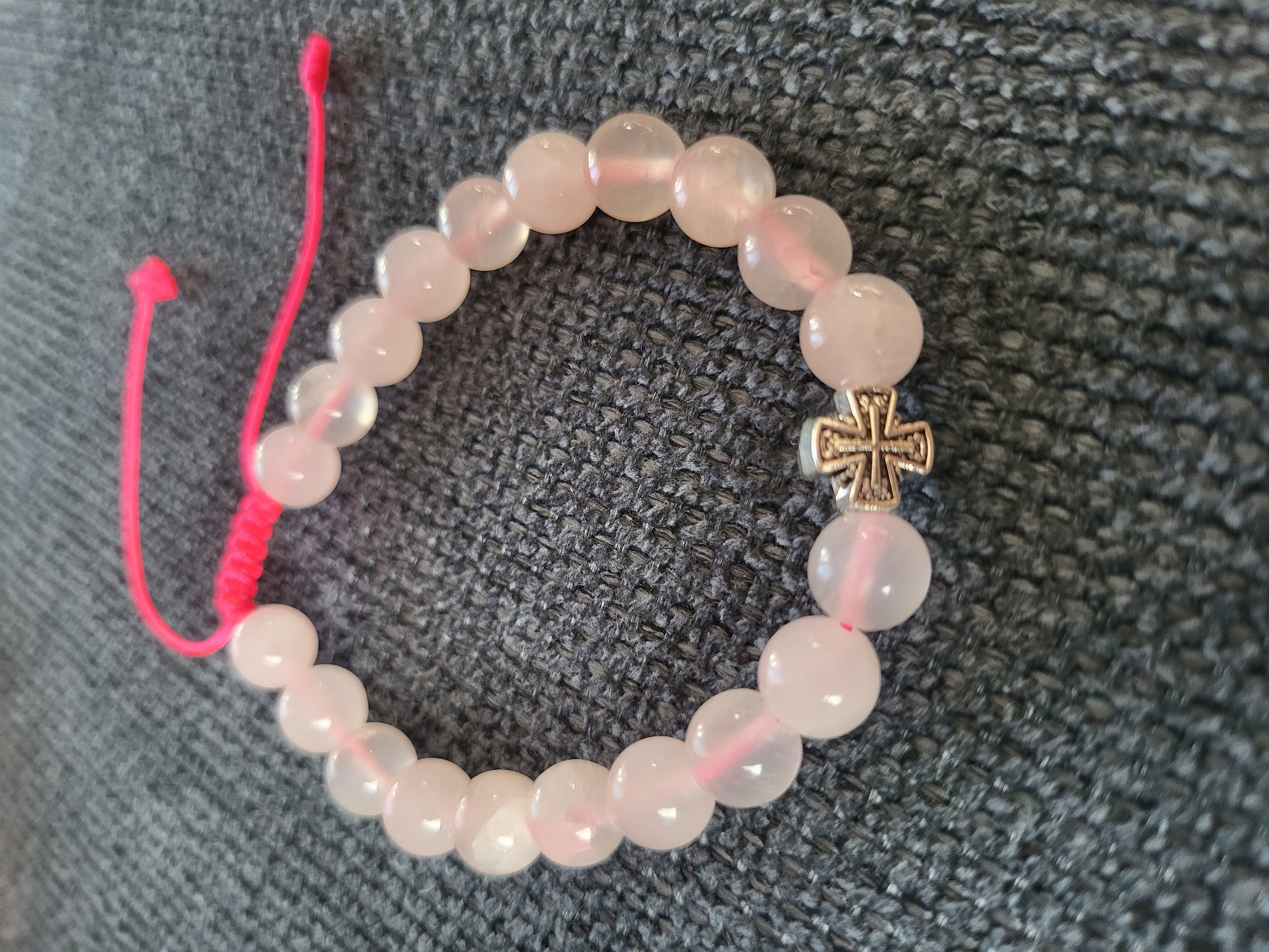 A close-up shot shows a pale pink beaded bracelet with a silver cross charm. The bracelet is made of smooth, rounded beads and is strung on a bright pink cord. The cord is tied at one end with a knot and has two loose ends extending from it. The bracelet is resting on a dark grey, textured surface, possibly a piece of fabric or upholstery. The lighting is bright, highlighting the translucent quality of the beads and the details of the cross charm. There are no people or animals visible in the image. The background is out of focus, making the bracelet the sole subject of attention. The location is not specified in the image.