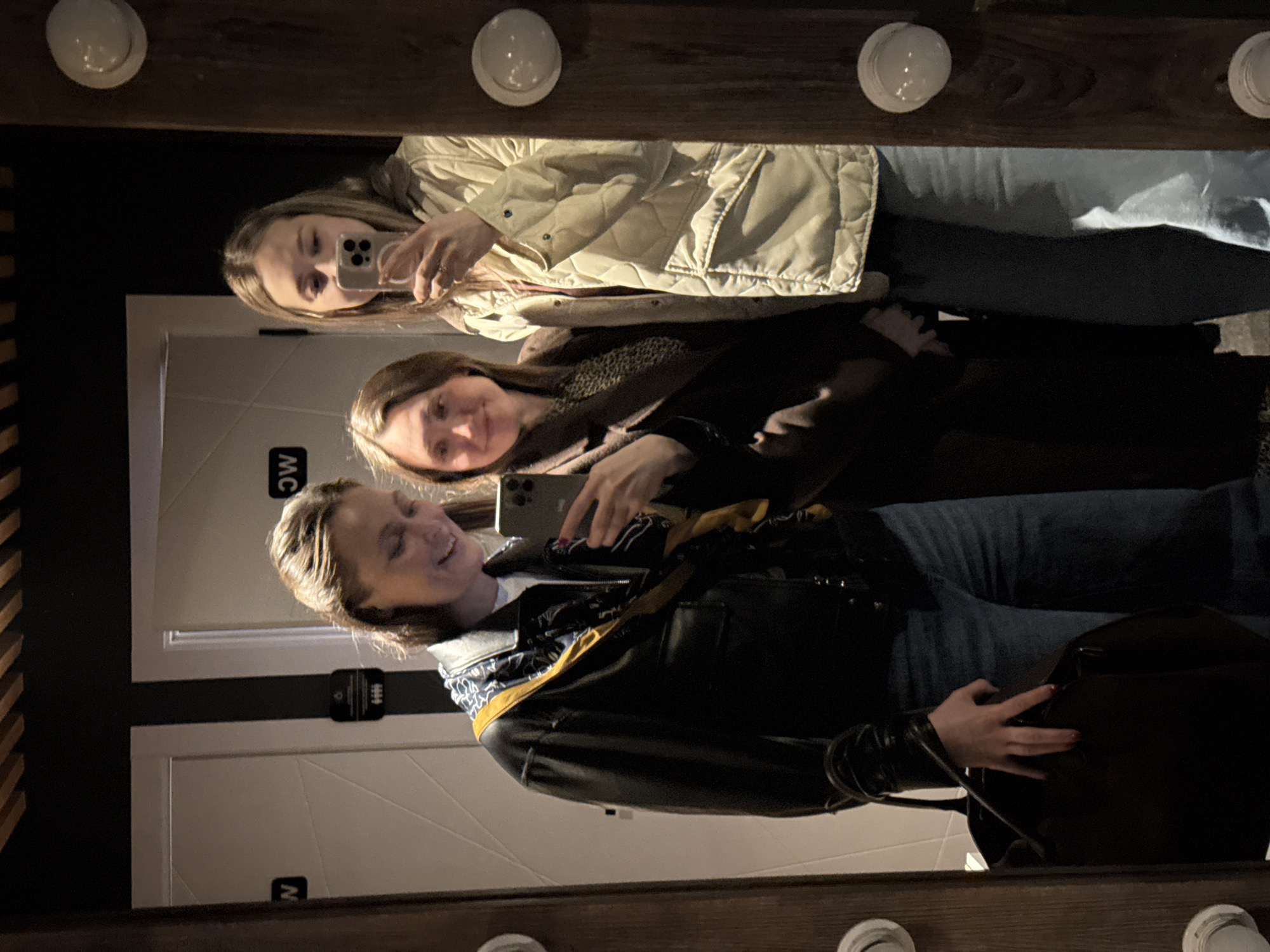 Three women are taking a selfie in a mirror. The mirror has light bulbs around the perimeter. The women are standing in front of bathroom doors with men's and women's symbols on them. The women are holding their phones. It is night. The city of Chaikovsky, Russia is not visible.