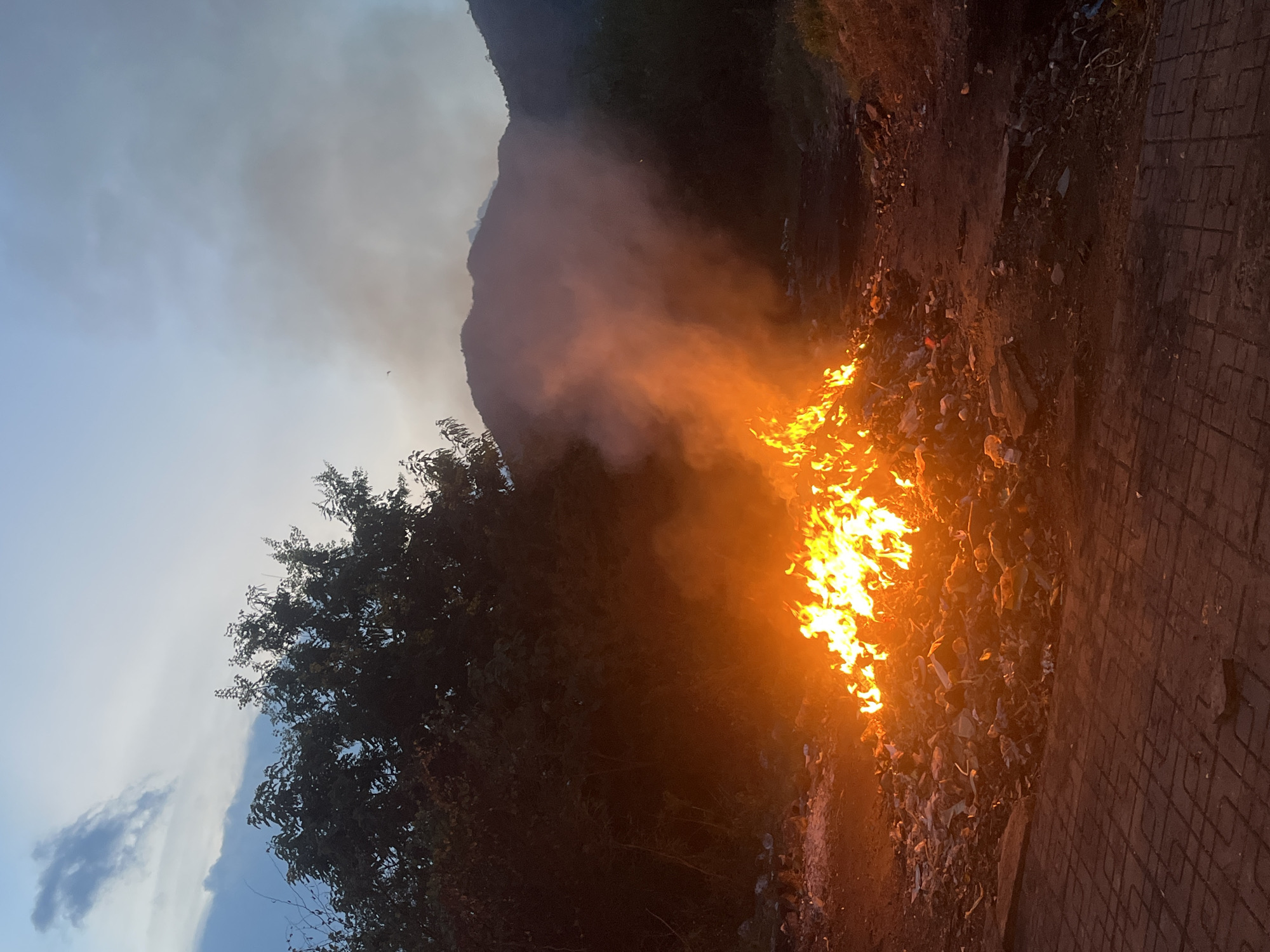 A large pile of burning debris is located alongside a road in Bac Nha Trang Ward, Vietnam. Flames rise from the ground, emitting thick smoke. Vegetation is present on the left side of the frame, with mountains visible in the background. A patterned brick surface covers part of the road on the right. The sky is overcast.