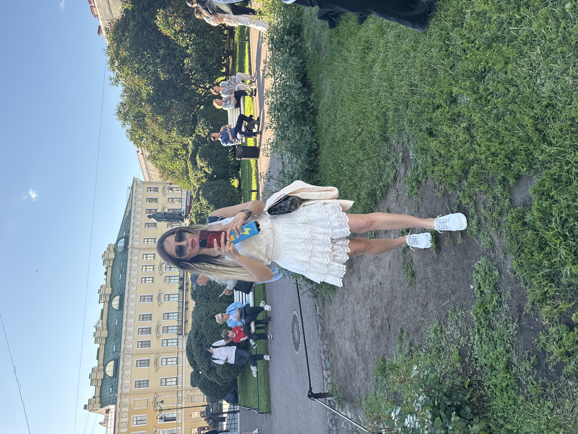 In this image, a young woman with blonde hair, wearing sunglasses and a white lace dress, is taking a selfie in an outdoor setting. She holds a red smartphone in her right hand, which is pointed towards herself. The woman is standing on a path, with a grassy area to her right and a building in the background. To her left, there are people sitting on benches and walking in a park. The building appears to be a historical yellow structure with green accents on its roof. The sky is clear and blue, suggesting it is daytime with good weather. The overall scene depicts a casual outing in a public space, possibly a park or a street lined with buildings.