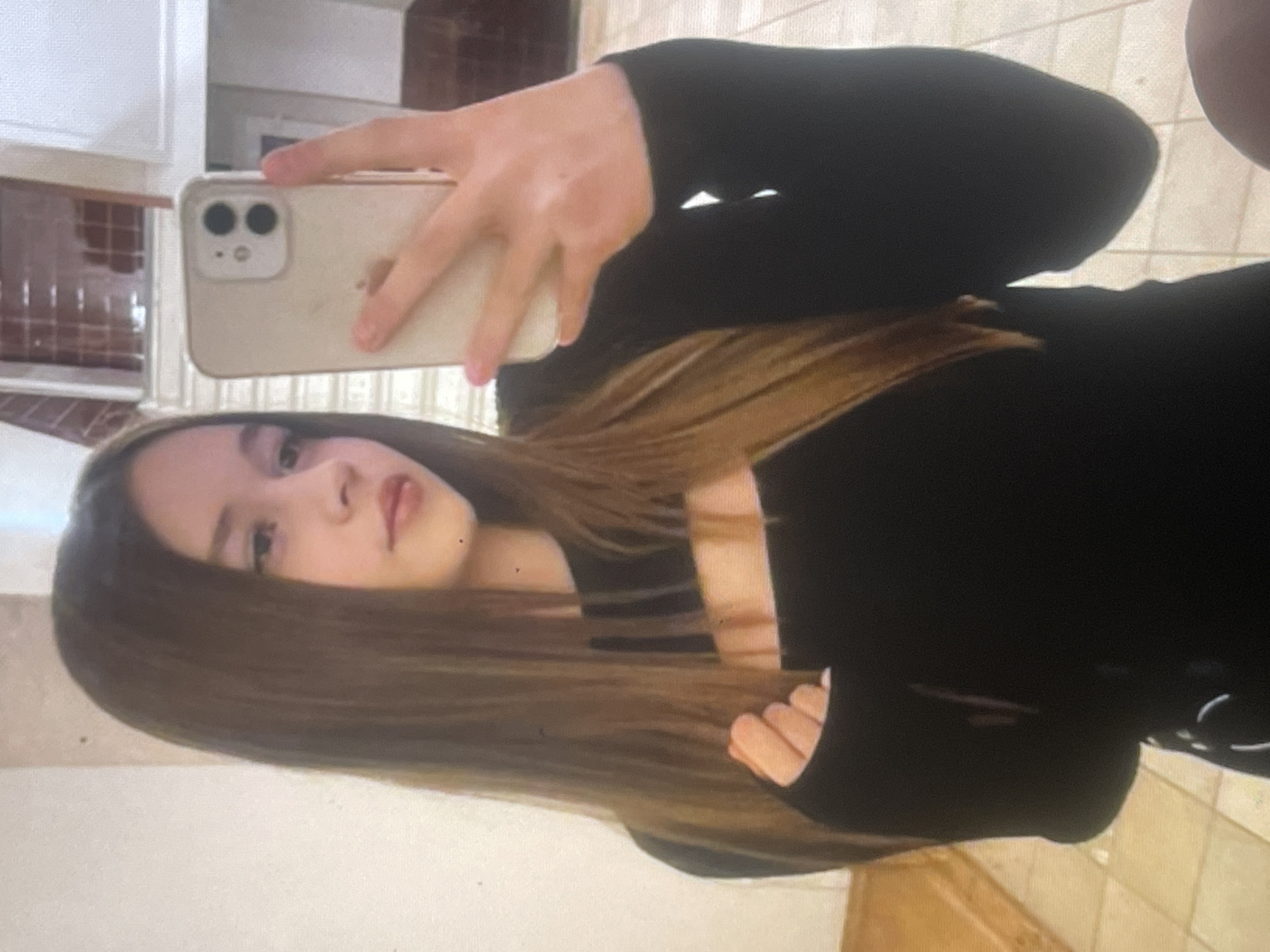 An image, rotated 90 degrees clockwise, captures a young person taking a mirror selfie indoors. The primary subject is a person, likely female, with long, straight brown hair, whose face is partially visible in the bottom left quadrant of the frame. She has a neutral expression. Her right hand, holding a light-colored smartphone (possibly an iPhone 12 or similar with its dual camera module), is prominent in the upper half of the image. She is wearing a black long-sleeved top, which appears to be a form-fitting garment.
The setting, visible in the mirror's reflection, appears to be a domestic interior. Elements in the background include white cabinetry or wall panels on the left and reddish-brown tiled surfaces, possibly a kitchen counter, backsplash, or floor, with a grid pattern. The lighting suggests an indoor environment, possibly with diffused natural light or artificial lighting. No specific time of day or weather conditions can be determined. There is no visible text in the image. The provided location context is Poltava, Ukraine.