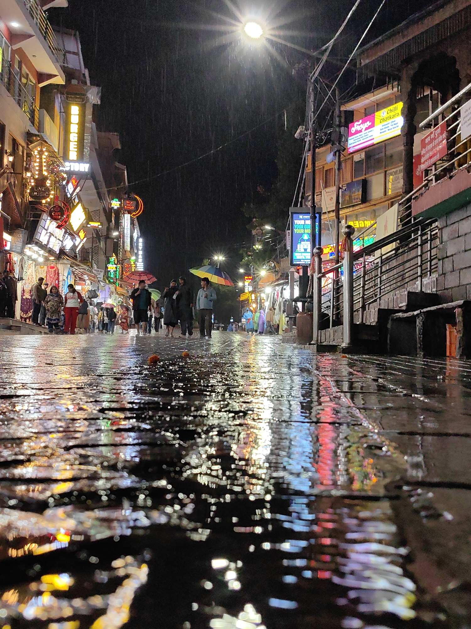 This image depicts a nighttime scene on Mall Road in Manali, India, during a rain shower. The street is wet and reflects the vibrant neon signs of various shops and restaurants lining the buildings. People are visible walking along the street, some holding umbrellas in an array of colors. The foreground is dominated by the wet pavement, with distorted reflections of the lights creating an impressionistic effect. The background shows the multi-story buildings with illuminated signage, indicative of a commercial district. The overall atmosphere is one of a bustling street scene adapted to inclement weather, with the rain adding a unique visual quality to the urban environment.