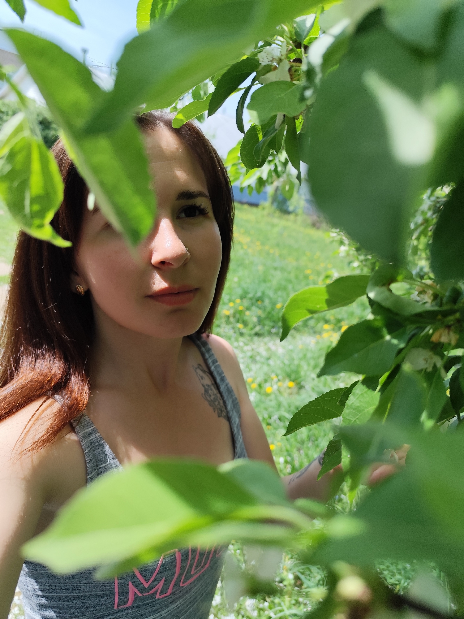 A woman is pictured from the chest up, framed by green foliage in the foreground and background. She has brown hair and is wearing a gray tank top. Her expression is neutral, and she has a nose ring in her left nostril. A tattoo is visible on her upper chest/shoulder. The background shows a grassy area with small yellow flowers and a glimpse of a light blue building. The photo seems to be taken outside during daylight. The image is not from Фоки, Russia.