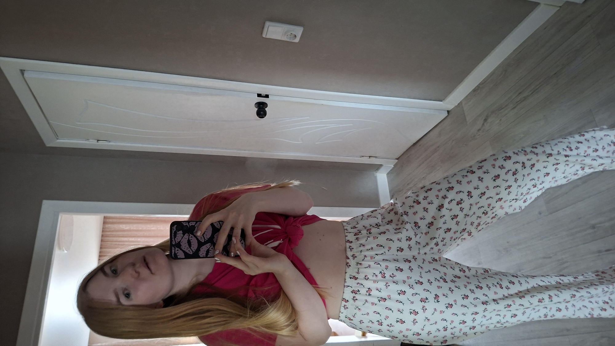 A young woman is captured in a full-body mirror selfie, standing in an indoor setting, likely a room or hallway in Chaikovsky, Russia. The image is presented rotated 90 degrees counter-clockwise.
The woman has long, light brown or blonde hair and a neutral to slightly contemplative expression. She holds a black smartphone with a distinctive pink or purple lips pattern on its case, using it to take the picture. A ring is visible on her left hand, and her fingernails appear to be painted a light color. She is wearing a bright pink or red short-sleeved crop top, which is tied at the front, exposing her midriff. Her lower body is covered by loose-fitting white pants patterned with small, repeating red floral designs.
The background features plain walls and a light-colored wooden or laminate floor. A white interior door with decorative grooves and a black handle is prominent behind her. Above the door frame, a white electrical outlet is mounted on the wall. The lighting suggests it is daytime. No discernible text is visible in the image.