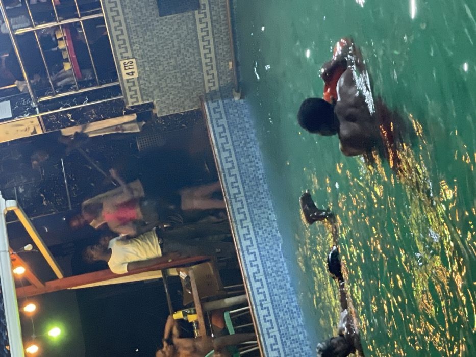 A group of people gathers near a pool, some partially submerged in the water. One person is positioned in the water, using a red floatation device. The pool's edge is marked with "4 FTS". Adjacent to the pool, people are standing or seated near tables and chairs under string lights. The scene appears to be illuminated at night.
