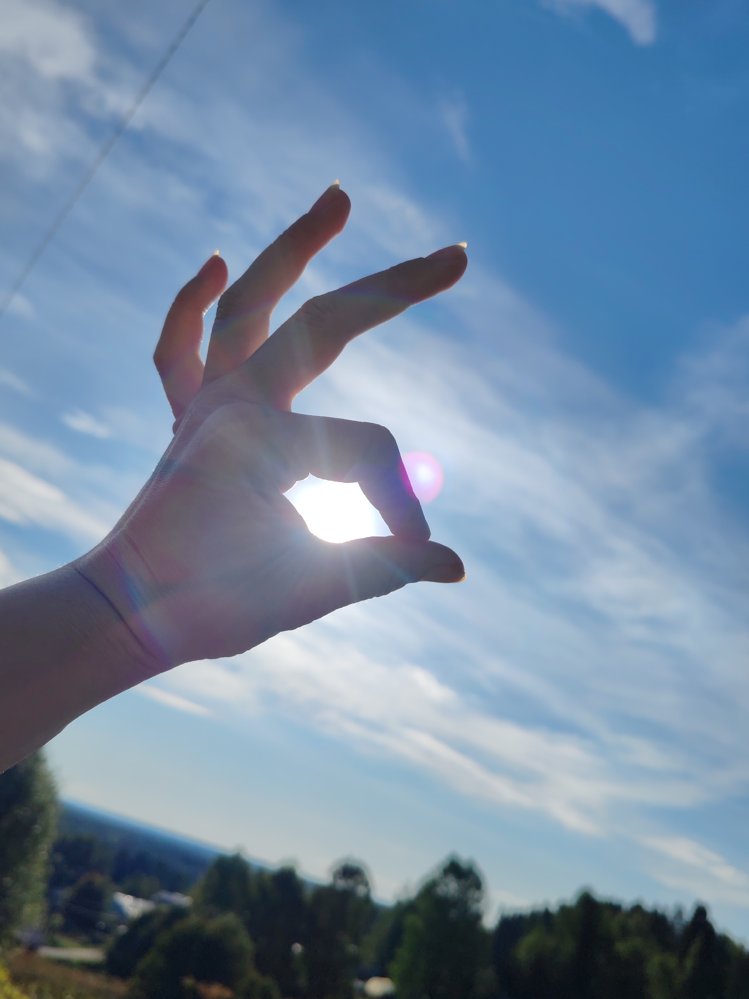A close-up, low-angle shot captures a human hand silhouetted against a bright blue sky with wispy clouds. The sun shines directly through the hand, which forms an "okay" symbol with its thumb and index finger, creating a lens flare effect with streaks of purple and yellow light. The background shows a blurry landscape of trees and distant buildings, suggesting an outdoor setting. This image was taken in Chukhlem, Russia.