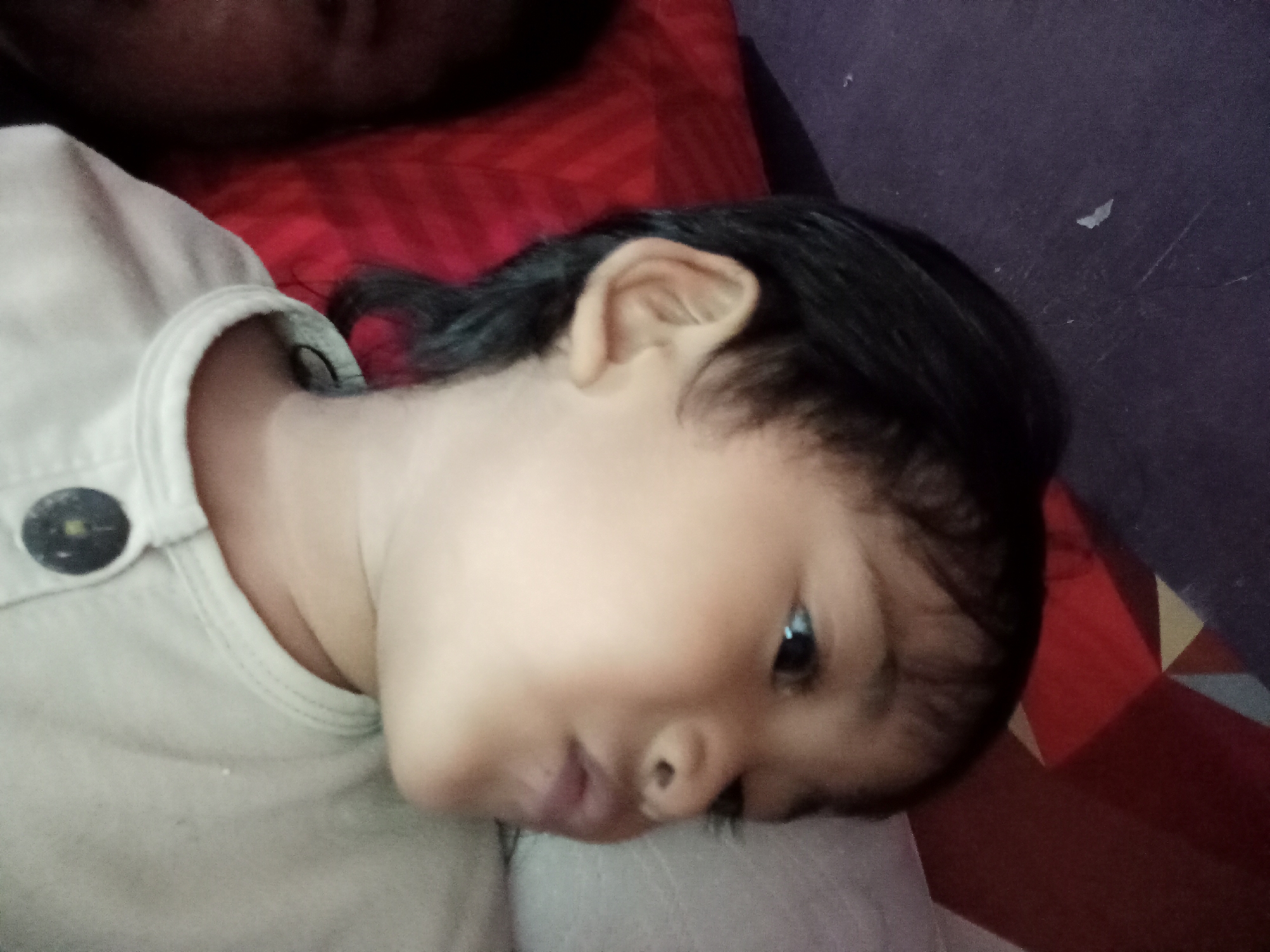 The image shows a close-up, low-angle view of a young child's face and upper body, lying down. The child has dark hair and is wearing a light-colored shirt with a button. The child's head is tilted to the right, with their left eye visible. The background consists of a red and patterned surface, and a darker, possibly purple, wall or surface. The lighting suggests an indoor setting. The provided metadata does not include information about the specific location within Bloro, Indonesia, or the date and time the photograph was taken.