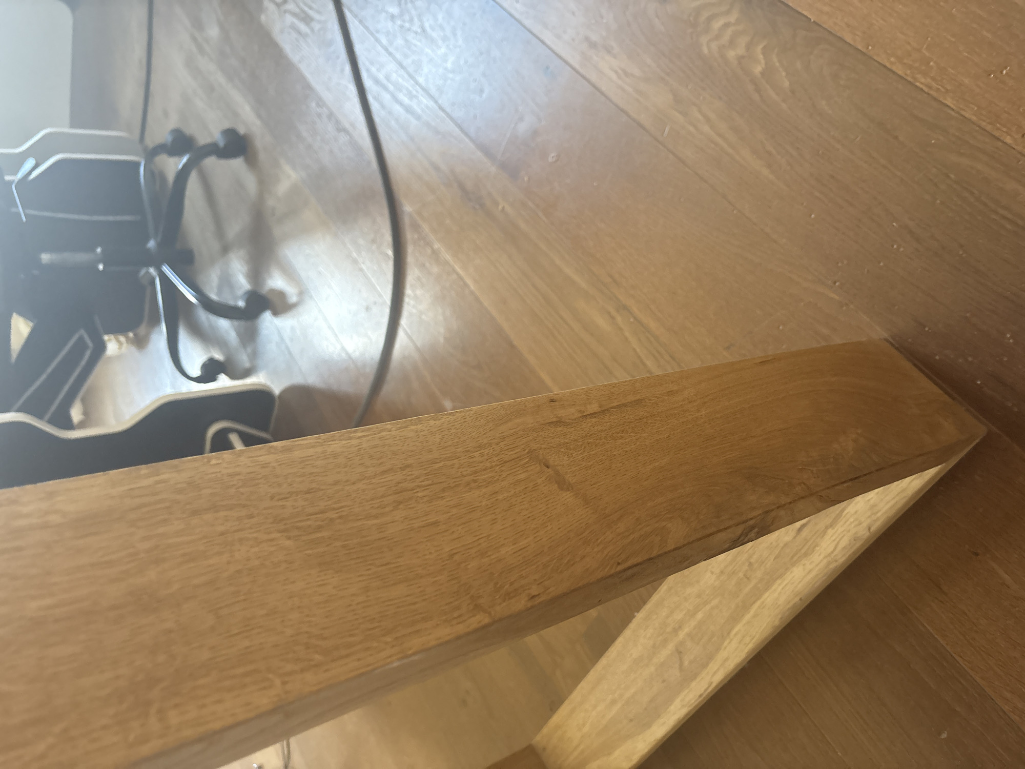 The image shows a close-up view of a wooden table leg and the wooden floor in front of it. In the background, out of focus, is a black office chair with wheels. A black cable runs across the floor. The scene appears to be indoors, likely in a residential or office setting. The location in the city of Orekhovka, Russia is not discernible from the visual information provided in the image.