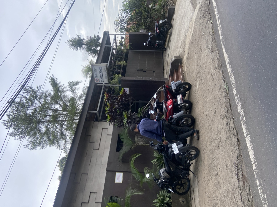 The image shows a man standing in front of a building in Cipatat, Indonesia, located at latitude -6.787 and longitude 107.434. The building is made of concrete blocks and has a brown wooden door. There are three motorcycles parked in front of the building. The man is wearing a blue shirt and black pants. He is looking at the motorcycles. The motorcycles are all black and have a red license plate. The motorcycles are parked on a gravel driveway. The driveway is lined with green plants and a few trees. The driveway leads up to the building, which has a sign above the door. The sign says "dr. MARISKA HARIS." The sky is overcast, and the background is blurred. The image was taken during the day. The man is not interacting with the motorcycles. There are no other people in the image.