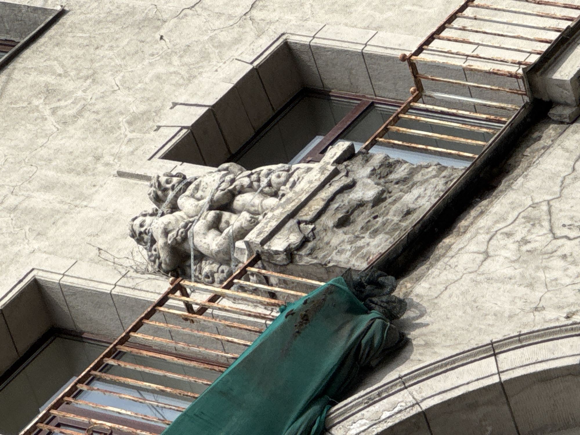 The image presents an upward-angled, close-up view of a weathered building facade, likely located in Saint Petersburg, Russia, based on context. The primary subject is an ornate stone carving centered beneath a deeply recessed window, flanked by a rusted fire escape.
The building's exterior is constructed from a light-colored material, possibly stucco or stone, characterized by a network of fine cracks indicating significant age. Two deeply set windows are visible, featuring dark glass and simple dark metal frames.
The central feature is a detailed stone sculpture depicting several cherubic or putti-like figures intertwined with drapery and a vertical element that resembles a cross or staff. The carving is weathered but retains clear definition, blending into the building's overall aged aesthetic.
To the right of the sculpture, a rusty metal fire escape or ladder runs diagonally downward across the facade. Its horizontal rungs and vertical supports are heavily corroded, showing distinct reddish-brown rust. Below the sculpture, a bundled mass of dark green fabric or tarp, possibly an old awning or protective covering, is draped over a part of the fire escape structure; it appears soiled or stained.
At the bottom right, a curved architectural element, potentially part of an arched window or decorative feature, is partially visible. The scene is illuminated by bright daylight, suggesting clear weather conditions and casting distinct shadows that highlight the textures of the stone and rusted metal. No visible text is present in the image.