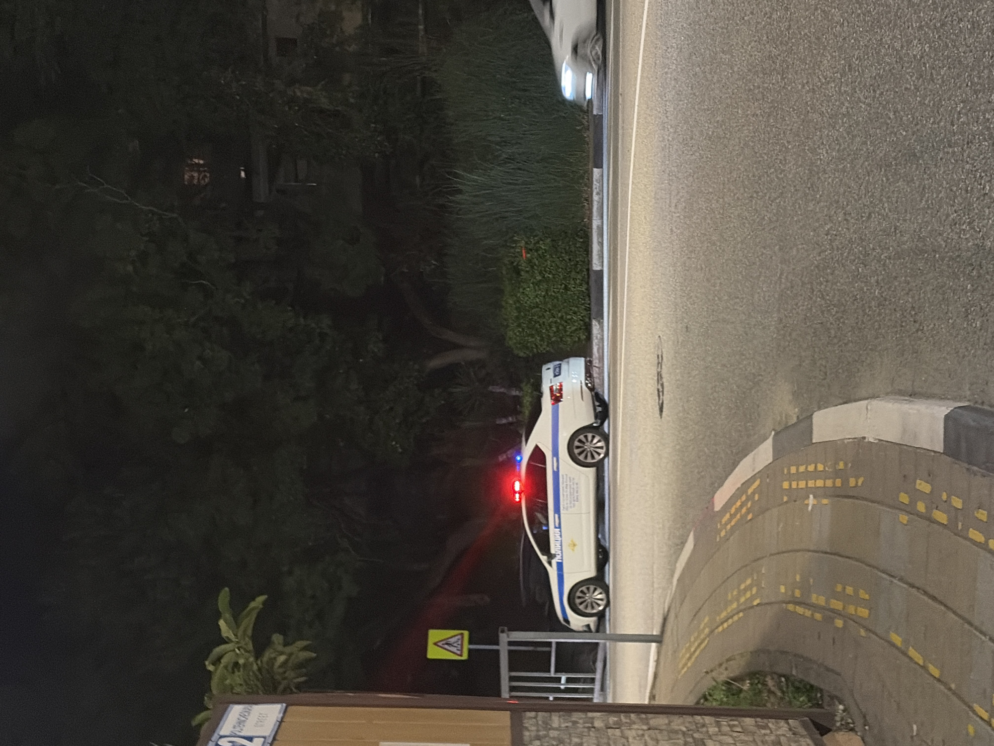At night on a street in Sochi, Russia, a white police car with its red and blue emergency lights activated is parked on the right side of the road. The car has the word "ПОЛИЦИЯ" (Police) written on its side in blue letters. A yellow warning sign with a black border and a black pedestrian symbol is visible on a pole to the left of the police car. The car is positioned next to a curb with yellow markings and a sidewalk. In the background, there are trees and a building with lights on. Another white car is partially visible further down the road. The scene suggests a roadside incident or police presence.