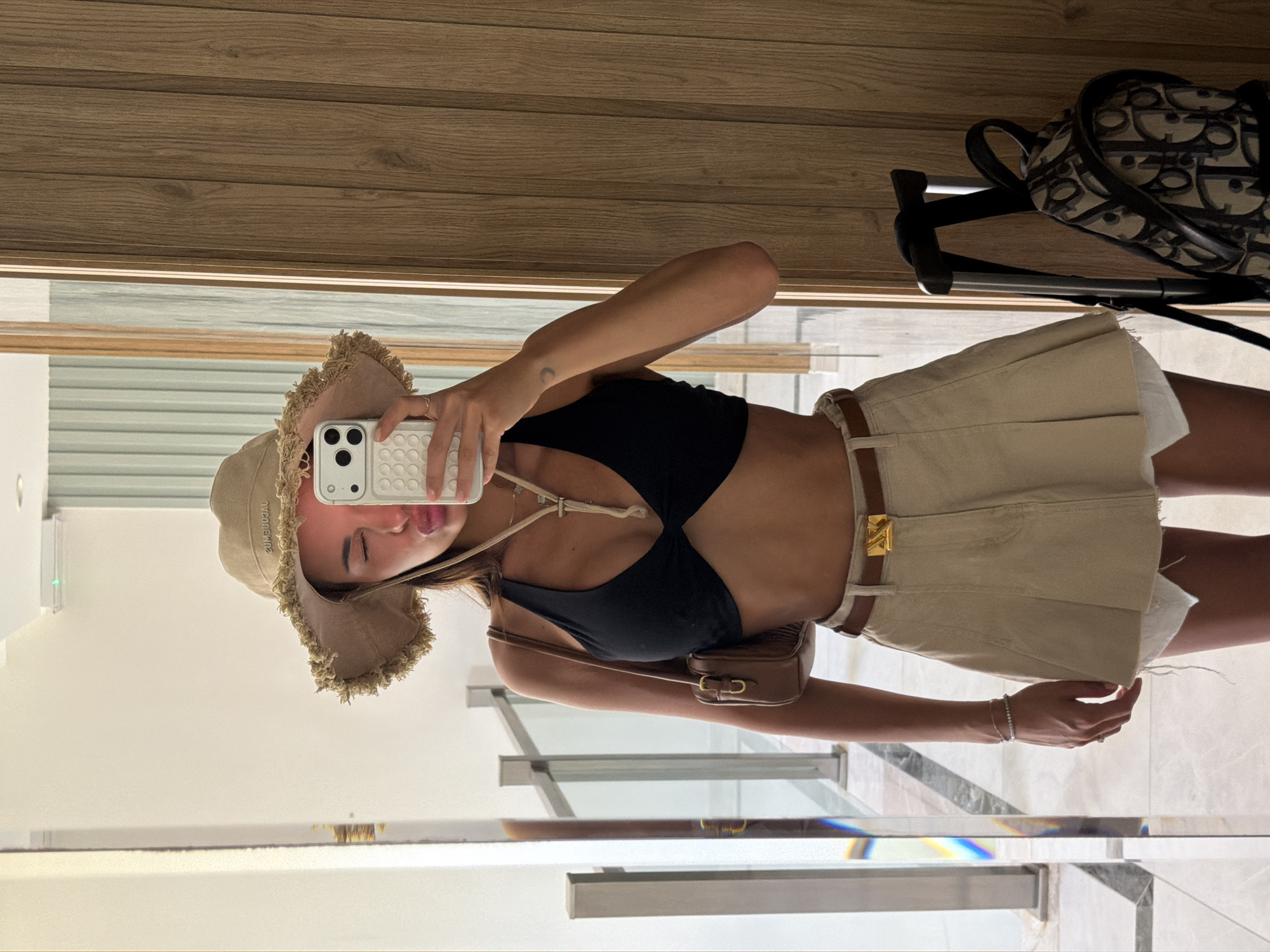 A young woman stands in a hotel hallway in Bodrum, Turkey, taking a mirror selfie. She is wearing a black bikini top, a tan bucket hat with frayed edges, and a tan pleated skirt with a white fringe hem and a brown belt with a gold buckle. She is holding a white iPhone in her right hand and has a small brown shoulder bag. A black suitcase with a patterned backpack attached to it is visible to her right. The hallway features a reflective floor and a wooden accent wall.