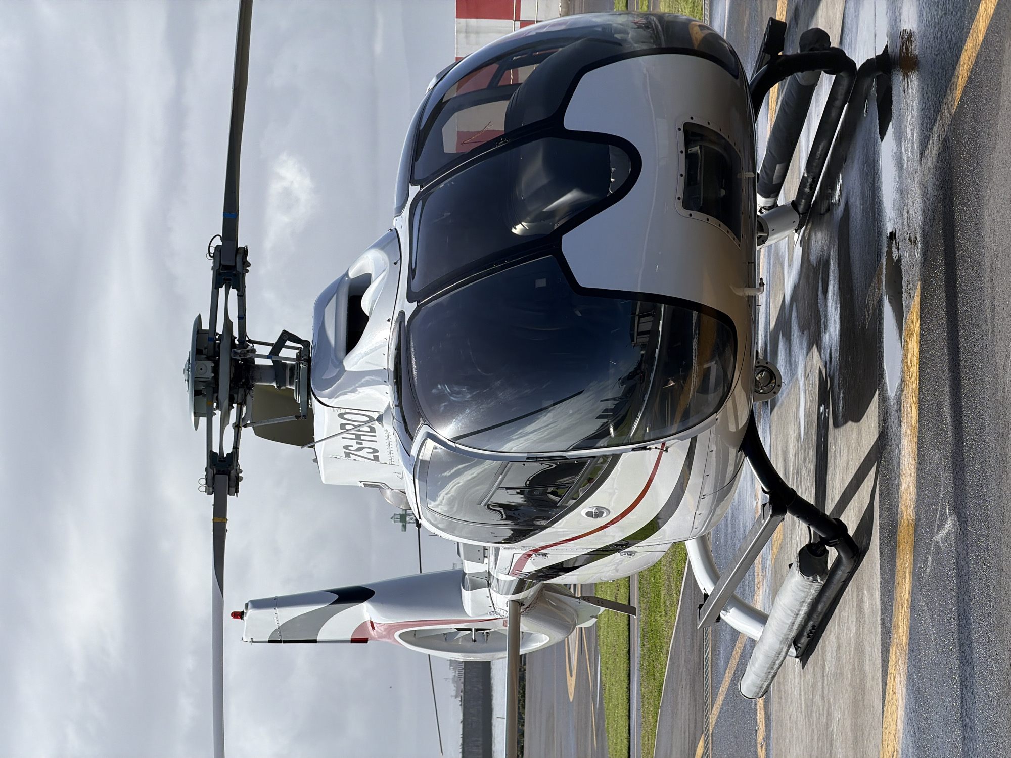 The image shows a white and black helicopter parked on a wet tarmac. The registration number "ZS-HBO" is visible on the tail. The ground is marked with yellow lines. The background includes a cloudy sky and distant buildings, suggestive of an airport environment. The image was taken in Cape Town, South Africa.