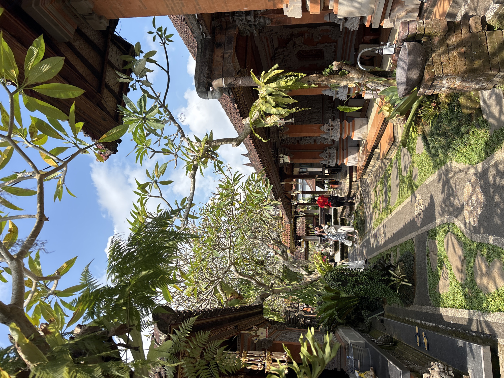 In Ubud District, Indonesia, a sunny day unfolds with lush greenery and traditional Balinese architecture. The scene captures a courtyard with a pathway lined with vibrant green grass and strategically placed stepping stones. On the right, an ornate stone sink with a faucet stands atop a brick pedestal, surrounded by tropical plants. Adjacent to it, an intricately decorated building with terracotta roofs and detailed carvings features an open-air walkway. Two women, one in a light-colored dress and the other in a patterned outfit, are walking away from the viewer on the pathway, engaged in conversation. Further down the path, a person in a red shirt is also walking. Lush trees with leafy branches extend from the foreground, their leaves creating dappled shadows on the ground. The overall atmosphere is serene and picturesque, showcasing the typical beauty of Ubud.