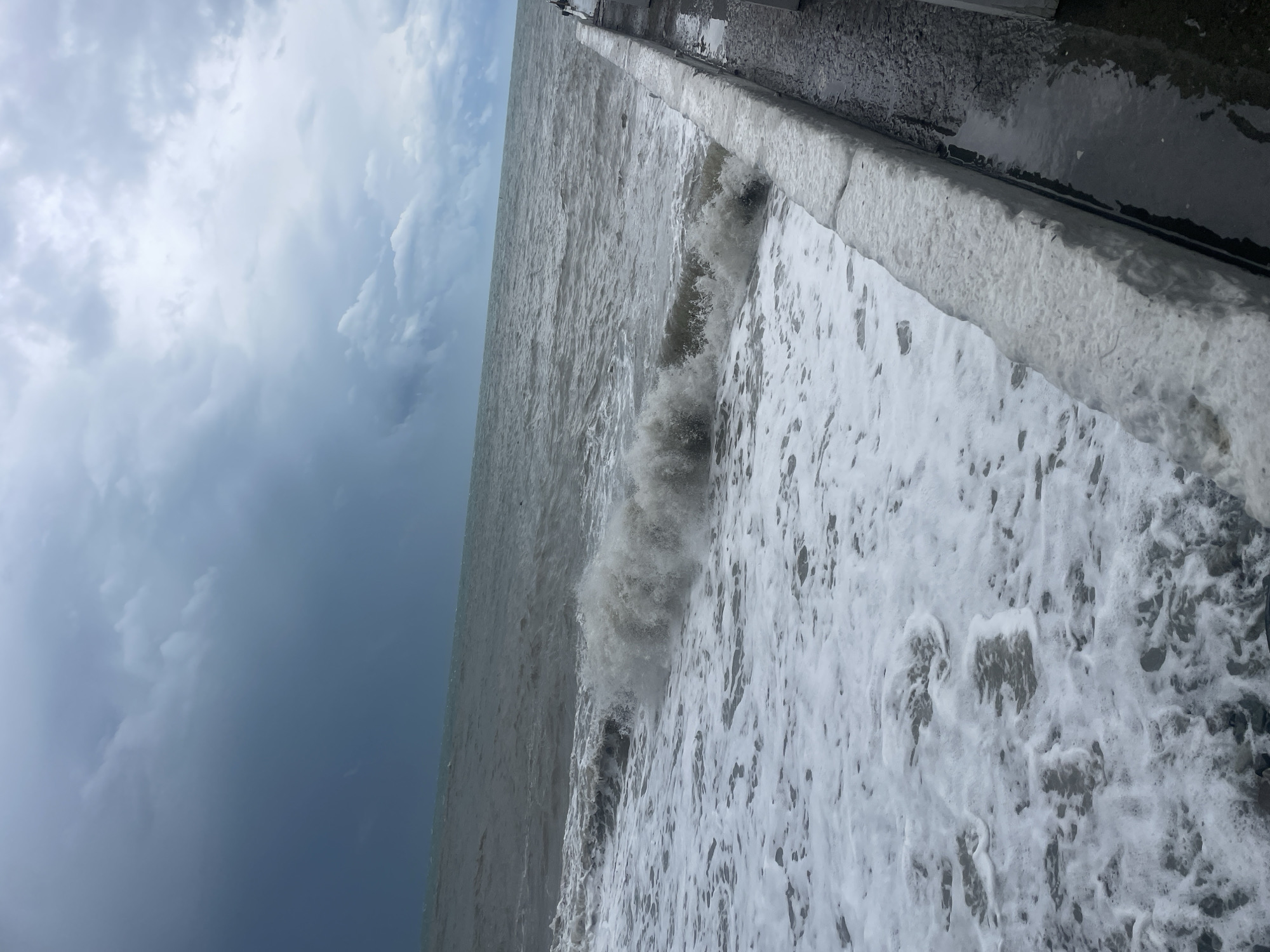 The image shows a stormy day at the Black Sea in Sochi, Russia. Heavy waves crash against the embankment, and the sky is filled with dark storm clouds. The perspective is from the embankment, looking out to the sea and up at the sky. There are no people visible.
