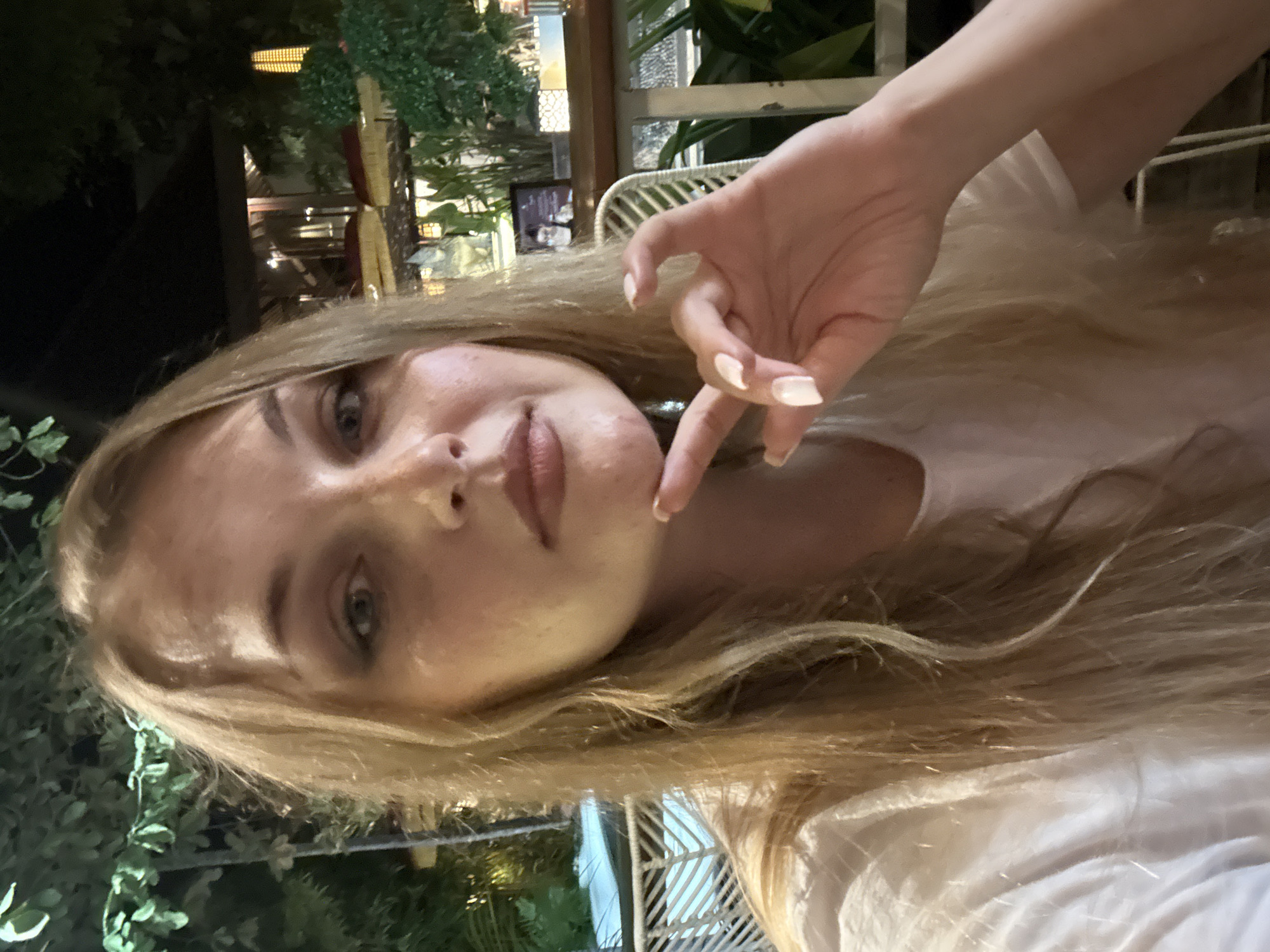 A close-up, selfie-style photograph depicts a young woman with blonde hair and fair skin, wearing a white t-shirt. She is indoors, with a blurred background that suggests an outdoor seating area, possibly at a restaurant or cafe in Denpasar, Indonesia. The environment features lush greenery, a dark, thatched roof structure, and outdoor seating with white wicker chairs and tables. The lighting is dim, indicating it is evening or nighttime, with artificial lights casting a warm glow. The woman is looking directly at the camera, with her left hand resting on her chin, her fingers delicately arranged. Her expression is neutral. The overall ambiance suggests a casual, relaxed setting in an urban environment of Denpasar.