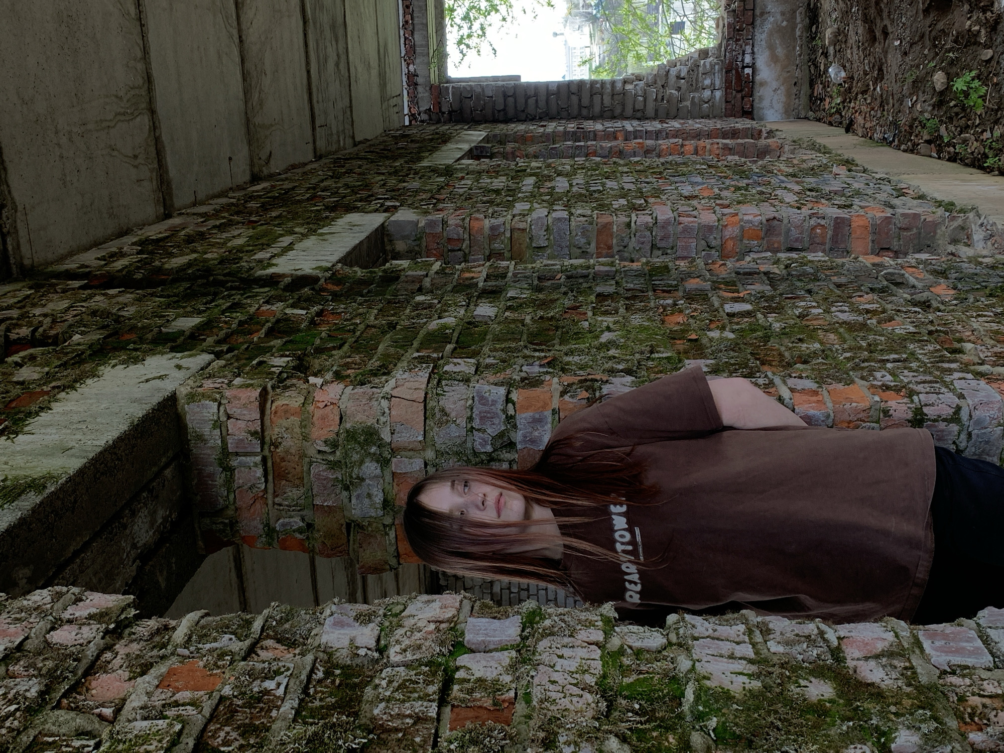 A person lies on a decaying brick structure within a building in Фоки, Russia. The structure shows significant wear, with moss covering many of the bricks. The person wears a brown shirt with white writing on it and dark pants. The building is dilapidated, with exposed concrete and brickwork. Above the brick structure, an opening reveals the city beyond.