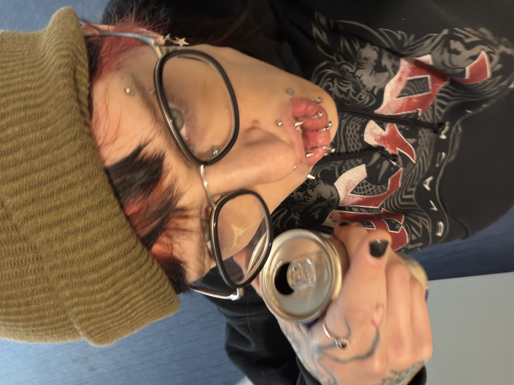 A person with multiple piercings in their ear and a nose piercing is shown holding an open aluminum can. They are wearing glasses and a knit cap. The person is wearing a black hooded sweatshirt with a large graphic. The person's left hand is visible, with dark nail polish, tattoos, and a ring. The floor is visible in the lower portion of the frame. The location is not identifiable as Sofia, Bulgaria from the available details.