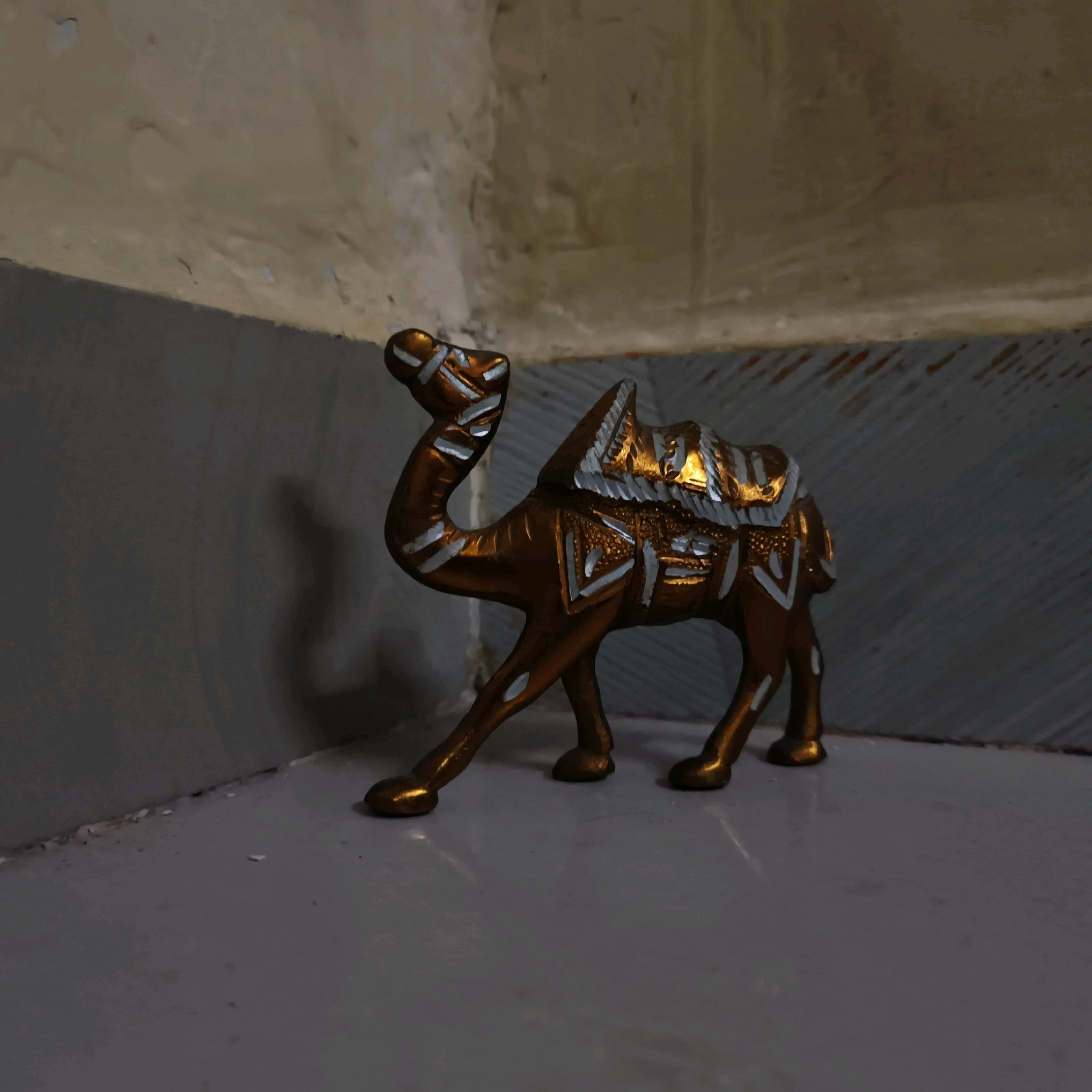 A close-up, low-angle shot shows a small, metallic, camel-shaped figurine. The figurine is predominantly bronze-colored with decorative silver accents, featuring a hump and intricate patterns along its body and saddle. It stands on a flat, light-colored surface, possibly a table or shelf, in a corner where two walls meet. The background consists of textured walls, one a muted beige with visible imperfections, and the other a darker grey. The lighting appears to be artificial, casting shadows and highlighting the metallic sheen of the figurine. The scene suggests an indoor setting, possibly a decorative display or a collectible item. There is no indication of Mumbai, India, or any specific city.