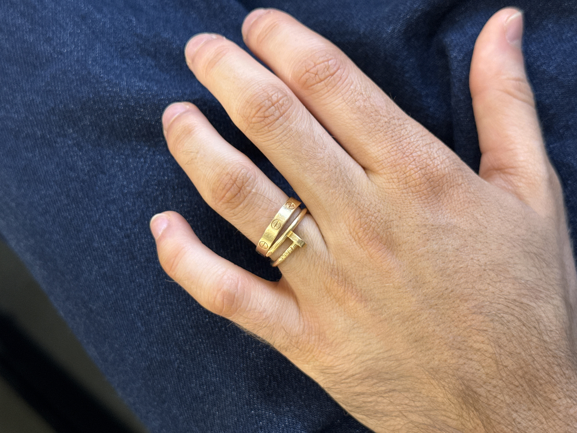 A close-up shot shows a person's hand resting on blue denim fabric, likely jeans. The hand has two gold rings on the ring finger. One ring is a wide band with regularly spaced circular indentations. The other ring is shaped like a nail, bent to form a ring shape. The image provides no information about the location, so Montreal, Canada, cannot be confirmed.