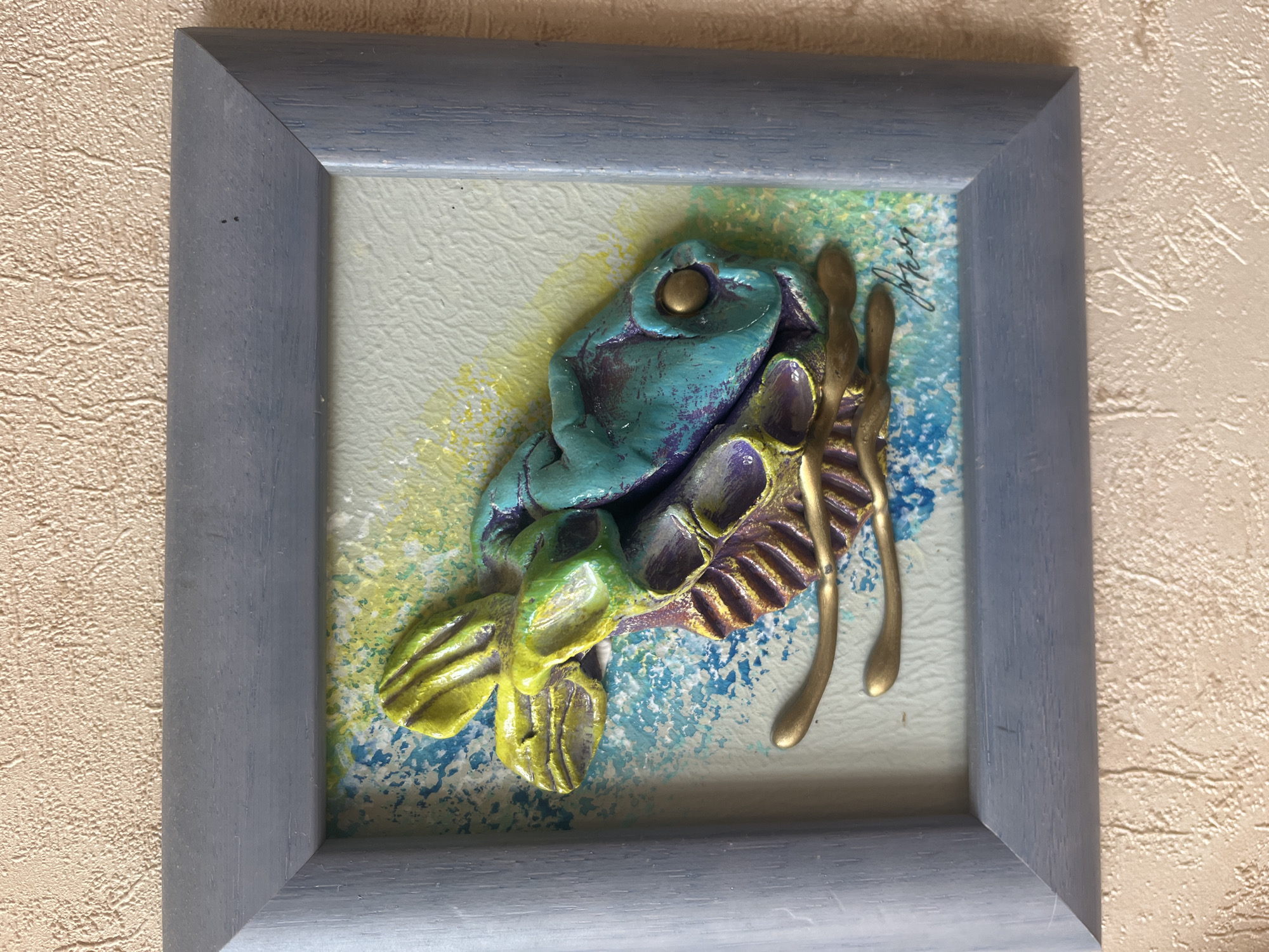 The image shows a framed 3D artwork with a textured background. The artwork itself is a colorful, sculptural representation of what appears to be a stylized animal head, possibly a horse or a dragon, with prominent features like a snout, ears, and an eye. It is painted in vibrant colors, including teal, purple, yellow, and green, with gold accents on what seem to be reins or a harness. The background of the artwork is a textured surface with splashes of blue and yellow paint, suggesting a dynamic or watery environment. The frame is a simple, light blue-grey wooden frame. The artwork is hanging on a textured, beige wall, consistent with interior decor. There is no information about the city of Vladimirovka, Russia, present in the image or its immediate context.