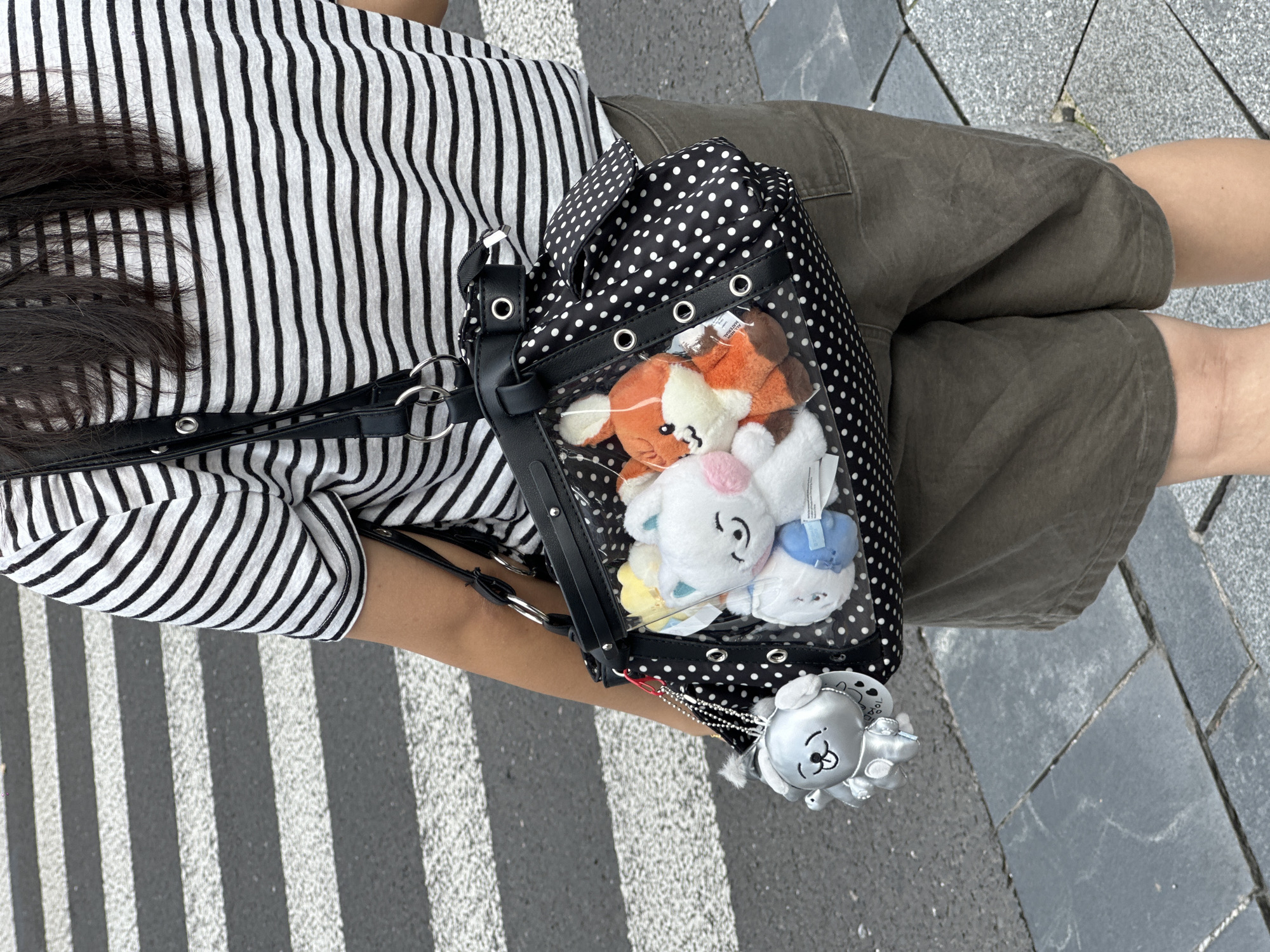 A person wearing a black and white horizontally striped t-shirt and brown shorts stands on a city street, partially obscuring a pedestrian crossing. The person is carrying a black and white polka dot bag with clear plastic panels, revealing several stuffed toys inside, including a white bunny and an orange fox. A silver dog-shaped keychain dangles from the bag. The ground is paved with grey stones and asphalt. The image appears to be taken in a public urban area, likely a street in Shanghai, China, given the context of the prompt.