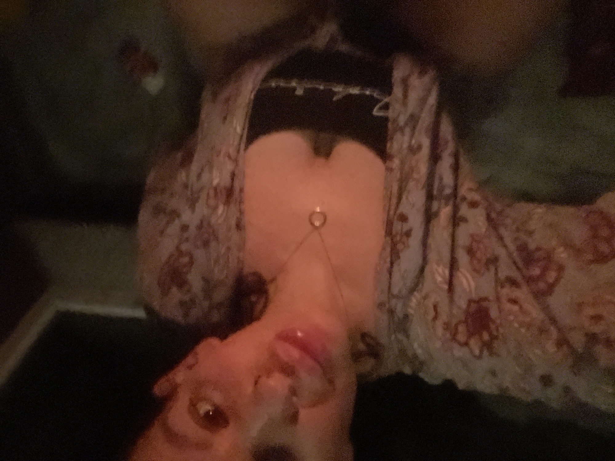 The image is a close-up, poorly lit selfie of a person. The subject is wearing a patterned open robe over a dark top. A necklace with a round pendant is visible. The person's face is partially obscured, with their lips and tongue visible. The background is dark and indistinct. No specific location in Plovdiv, Bulgaria is identifiable.