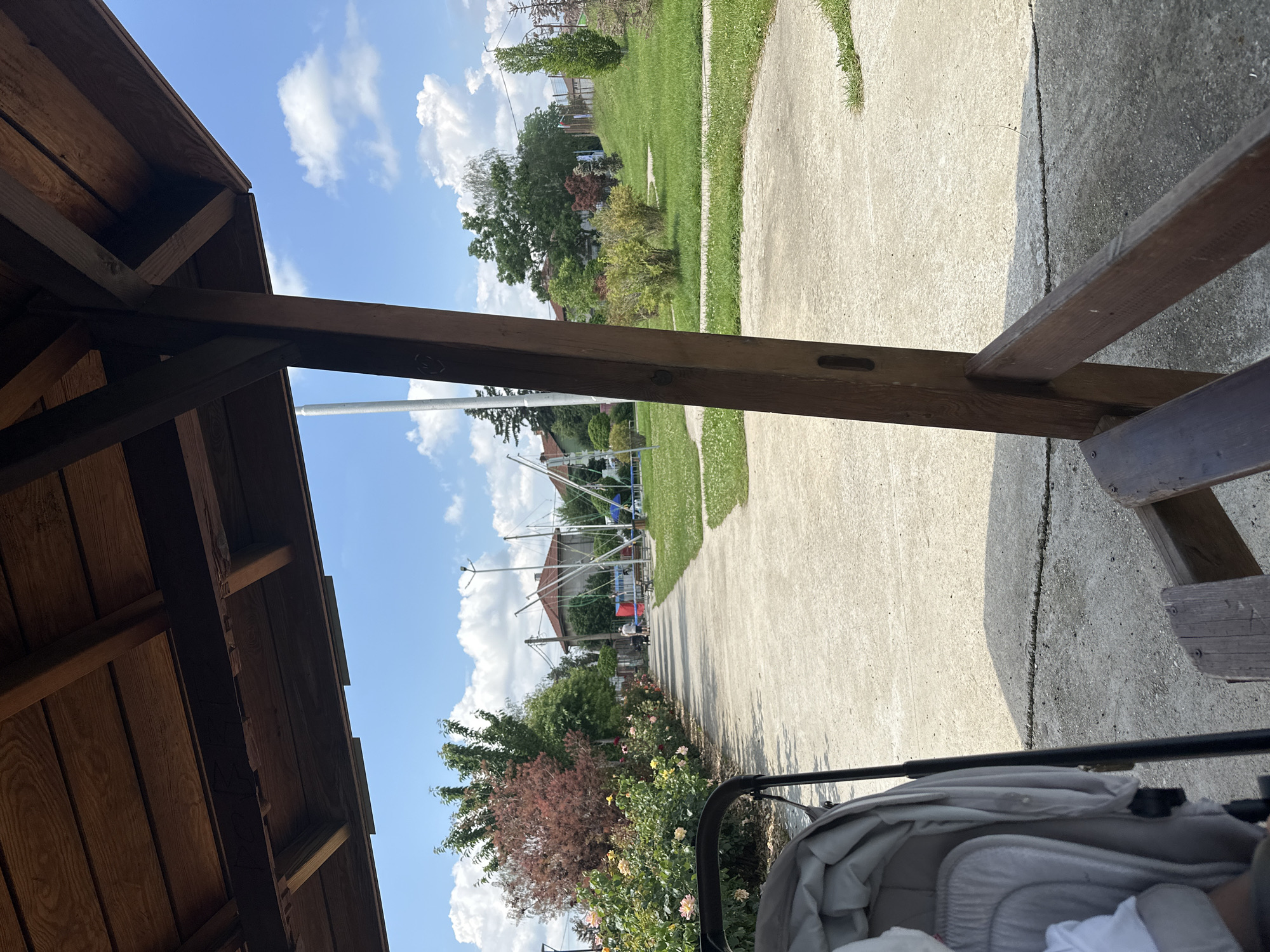 An eye-level view from beneath a dark brown wooden gazebo or pergola shows a paved path leading into a park or recreational area in Unknown, Unknown. The wooden overhead structure is visible in the upper portion, with a diagonal support beam on the right. In the lower left, the upper part of a light gray baby stroller is visible. The paved path, made of concrete, extends from the mid-left foreground into the background.
To the left of the path, a dense border of flowering bushes with pink, yellow, and red blooms lines the edge, followed by a large tree with reddish foliage and another with green leaves. In the mid-background, a person is walking away from the camera on the path. To the right of the path, a grassy area with scattered bushes is visible, and in the right mid-background, several tall metal structures with elastic cords, indicative of bungee trampolines, are present.
The background features residential houses with red-tiled roofs nestled among various trees. A tall white pole stands on the left side of the scene, and a double-headed street lamp pole is visible on the right. The sky is clear and bright blue with scattered white cumulus clouds, indicating a sunny day. Shadows are cast by the wooden structure onto the concrete in the foreground.