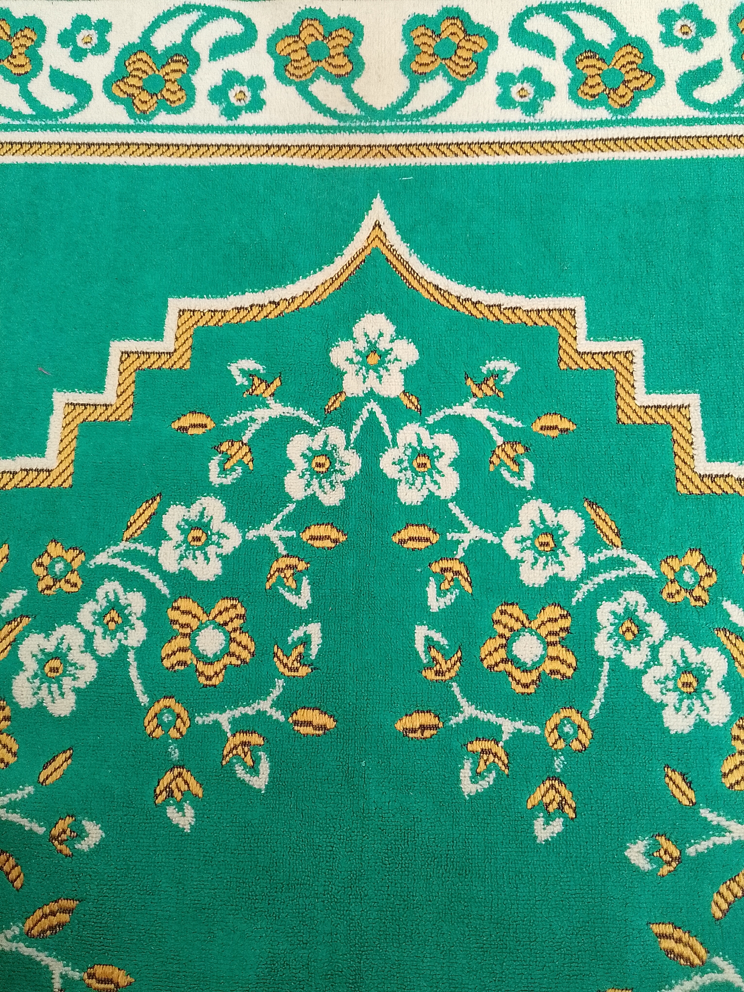 This is a close-up, high-angle, eye-level shot of a green prayer rug with a white and gold floral pattern. The rug is likely located in the city of Teep, Indonesia, although the exact location within Teep is not specified. The pattern features stylized flowers with white petals and yellow centers, interspersed with green leaves and small yellow floral accents. A decorative border of interlocking white and gold zigzags is visible at the top of the rug, above a band of white fabric adorned with more white and gold floral motifs. The texture of the rug appears to be soft and slightly fuzzy. There are no people, actions, or interactions depicted in the image. The environment is a flat, indoor surface.
