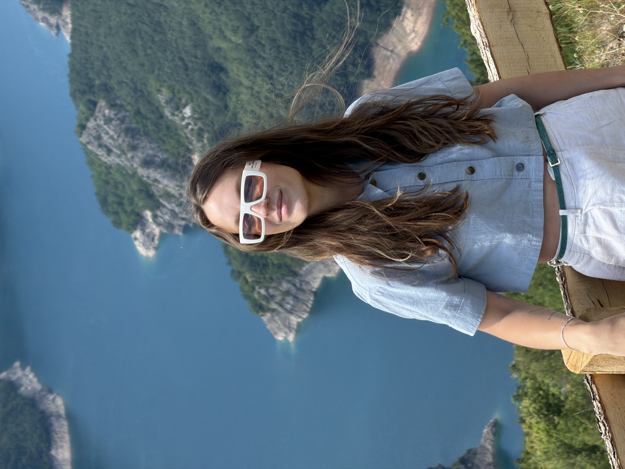 A young woman with long, brown hair and white-rimmed sunglasses stands in the foreground, looking to her left. She is wearing a light blue, cropped button-up shirt and white pants with a green belt. She has a bracelet on her left wrist. Behind her, a bright blue, winding body of water is surrounded by steep, tree-covered mountains. The scene appears to be a scenic overlook in Dubljevići, Montenegro.
