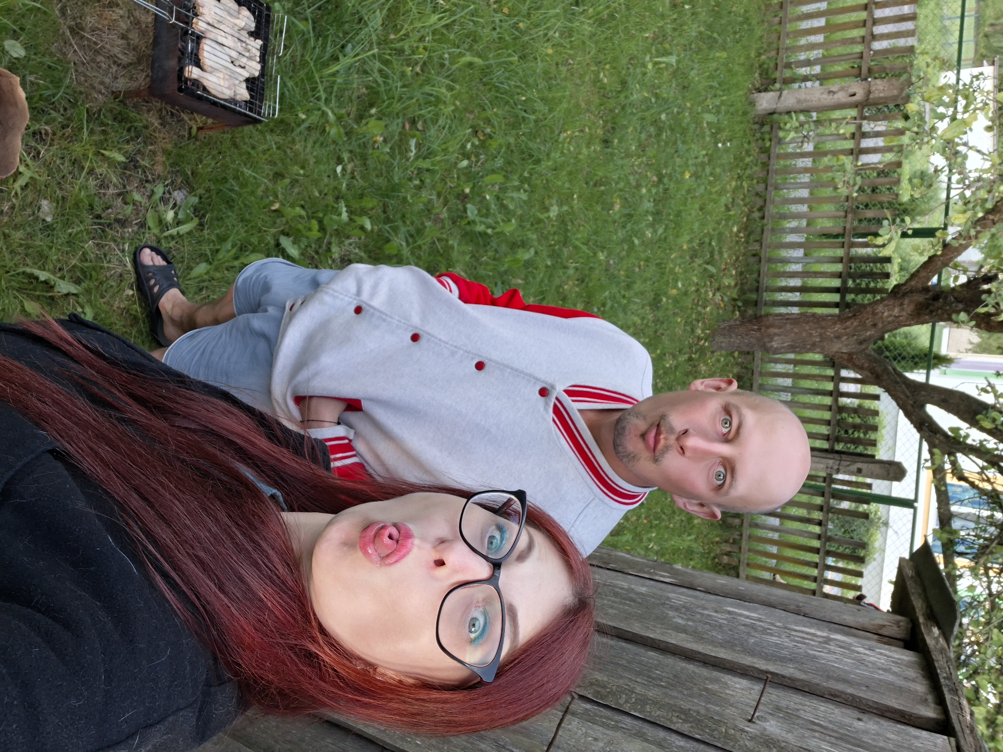 In an outdoor scene that appears to be the backyard of a residential property in Забалоцкі сельскі Савет, Belarus, a bald man with a goatee and a woman with long, red hair are taking a selfie. The man is wearing a light-colored bomber jacket with red trim and light blue shorts. He is puckering his lips and looking at the camera. The woman, wearing glasses and a black jacket, is sticking out her tongue and looking at the camera. Behind them, a wooden fence encloses a grassy yard. A small grill with raw meat on it is visible to the left of the frame. A rustic wooden structure is to the right of the couple. The overall impression is one of a casual, perhaps familial, outdoor gathering.