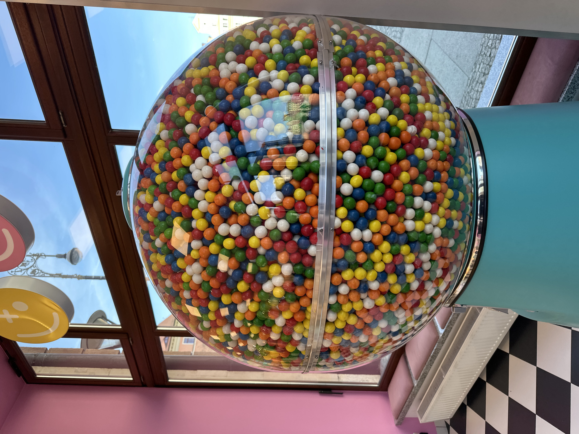 The image shows a large, spherical gumball machine filled with colorful gumballs. The machine is mostly clear, allowing the gumballs to be seen. It is mounted on a light blue cylindrical base. The background features a building, seen through a glass ceiling. A black and white checkered wall is visible on the right side. In the bottom left, a pink wall can be seen, along with a yellow smiley face decoration. The image was taken in Wrocław, Poland.