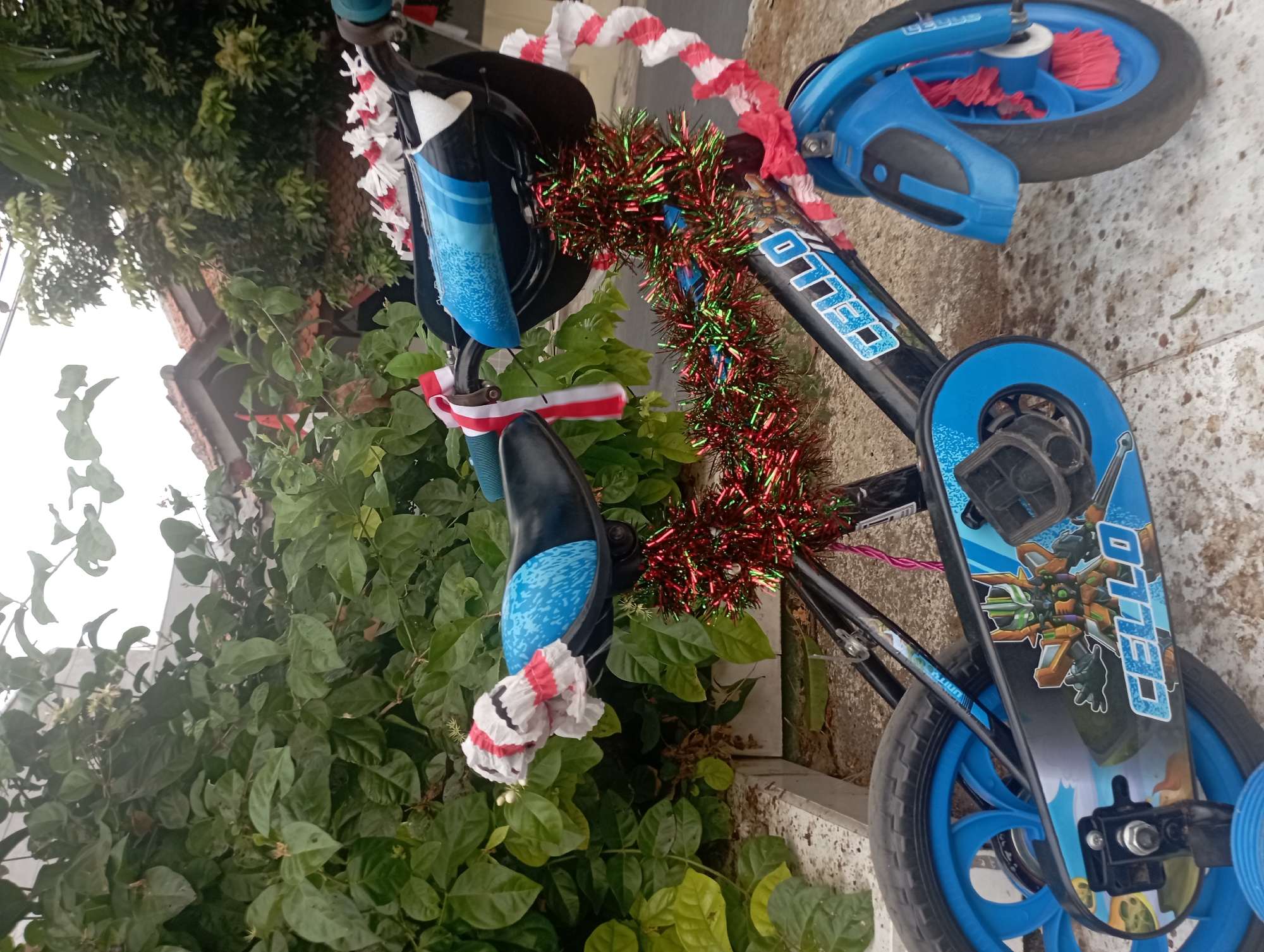 The image shows a close-up of a child's bicycle decorated with festive red and white streamers and green and red tinsel. The bicycle is black with blue accents on the wheels, fenders, and seat. The seat is decorated with a blue patterned fabric and a white and red streamer bundle. The handlebars are also adorned with red and white streamers. A green and red tinsel garland is wrapped around the frame of the bicycle, specifically around the handlebars and the front of the frame. The bicycle has a character sticker on the chain guard featuring a robot-like figure. The bicycle is parked on a tiled surface outdoors, with lush green foliage visible in the background. A residential building with a tiled roof is also partially visible behind the foliage. The scene suggests a celebratory occasion, possibly related to a local holiday or event in Purwakarta, Indonesia.