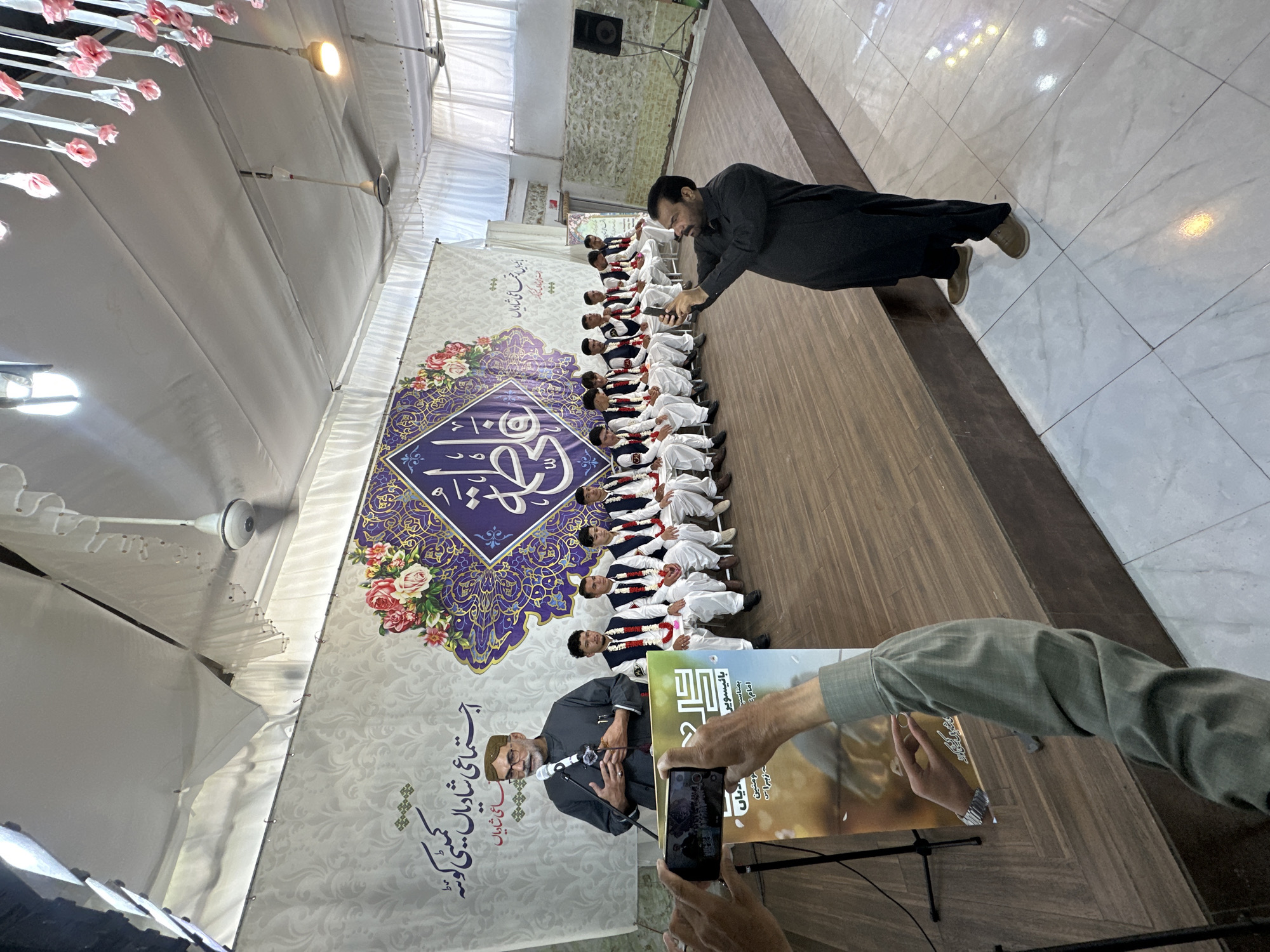 The image captures a gathering, possibly a wedding or cultural event, in what appears to be an indoor hall in Quetta City Tehsil, Pakistan. A line of individuals, likely young men, dressed in white attire with red sashes and dark vests, are seated against a decorated backdrop. A man in a dark gray robe is leaning over the group, possibly taking a photo with his mobile phone. Another man is standing in front of a poster that has been written in Urdu. The poster says "social wedding", "wedding committee in Quetta". The hall's interior is visible, with a decorated ceiling and patterned flooring. The general mood is festive and formal, suggesting a communal celebration.