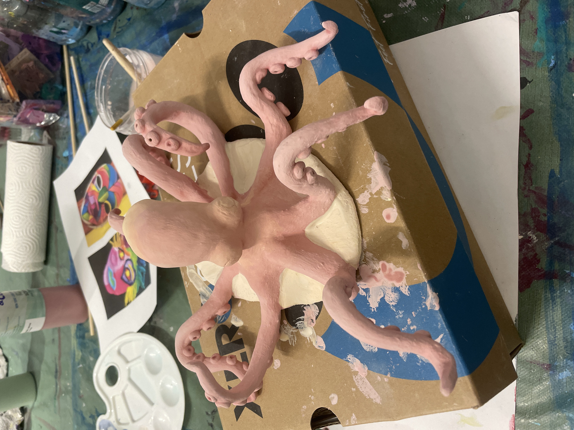 A handmade, pink clay octopus sculpture rests on a partially open cardboard box. The octopus is centered on the box, with its tentacles extending in various directions. The box has a brown base with blue and black graphic elements. The octopus is positioned on a light-colored base, potentially also clay, affixed to the box.
To the left of the box, on a painted surface that is likely a table, are painting supplies including: a tube of pink paint, a palette, a cup with paintbrushes, a roll of paper towels, and a printed image of a colorful abstract painting.
The image provides insufficient evidence to determine a specific location in Raanana, Israel.