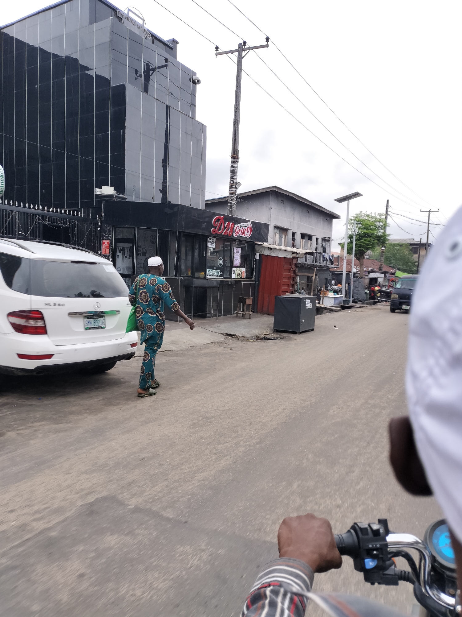 This media file depicts a daytime street scene in Alaiyabiagba, Nigeria, captured from the viewpoint of someone riding a motorcycle. The foreground shows the handlebar of the motorcycle, with a person's arm and a partially visible white-clad shoulder and head on the right.
In the mid-ground, a man dressed in a green and brown patterned traditional outfit and a white cap is walking along the asphalt road. He carries a green bag and is positioned next to a parked white Mercedes-Benz ML 350 SUV. The SUV's license plate, "SMW-832 JE," is clearly visible.
The background features a mix of buildings and commercial establishments. On the left, a multi-story black building with reflective glass panels rises, with the partial text "THE WORLD IS YOURS" visible near its top. Below it, a storefront displays a "DR Grill" sign and another sign indicating "OPEN MONDAY." A partially visible Heineken logo is on a fence. Further down the street, more modest gray buildings, various stalls, and street furniture, including electricity poles, power lines, and a solar-powered street light, line the road. A dark van is visible in the distance. The sky is overcast, suggesting a cloudy day.