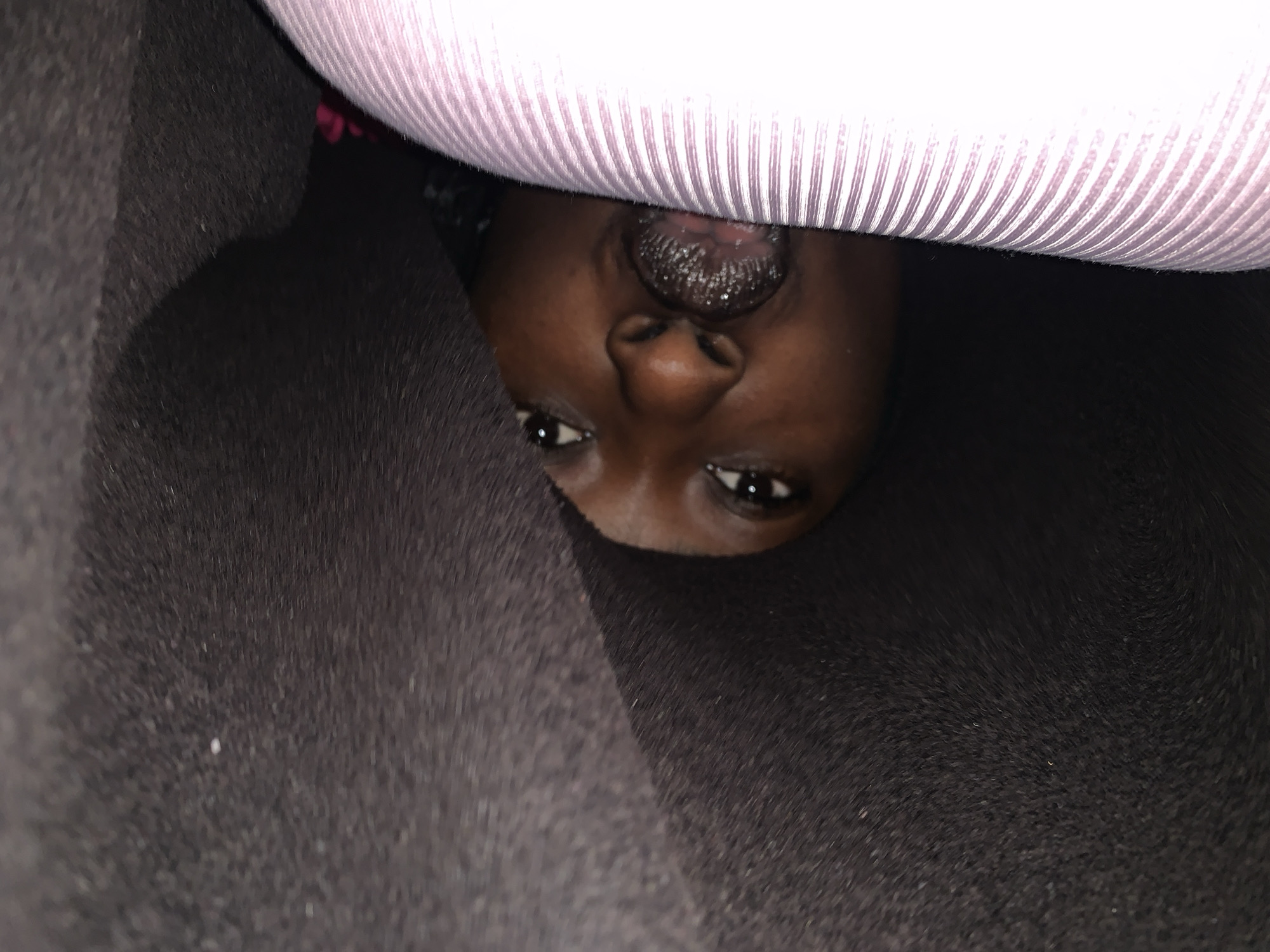 A close-up, low-angle, vertically oriented shot shows a young Black person's face emerging from between dark, textured material and a light pink, ribbed surface. The person's head is tilted, and they are looking directly at the camera. Their eyes are open, and their mouth is slightly agape, revealing their tongue which is coated with a dark substance, possibly lip gloss or balm. The dark material appears to be fabric, possibly the interior of a bag or a tightly wrapped blanket. The pink surface is ribbed and has a smooth, uniform texture, suggesting it could be a rolled-up yoga mat or a similar cylindrical object. The lighting is dim, casting shadows that obscure details in the background. No specific location in Jos, Nigeria, can be identified from the visual information provided in the image.