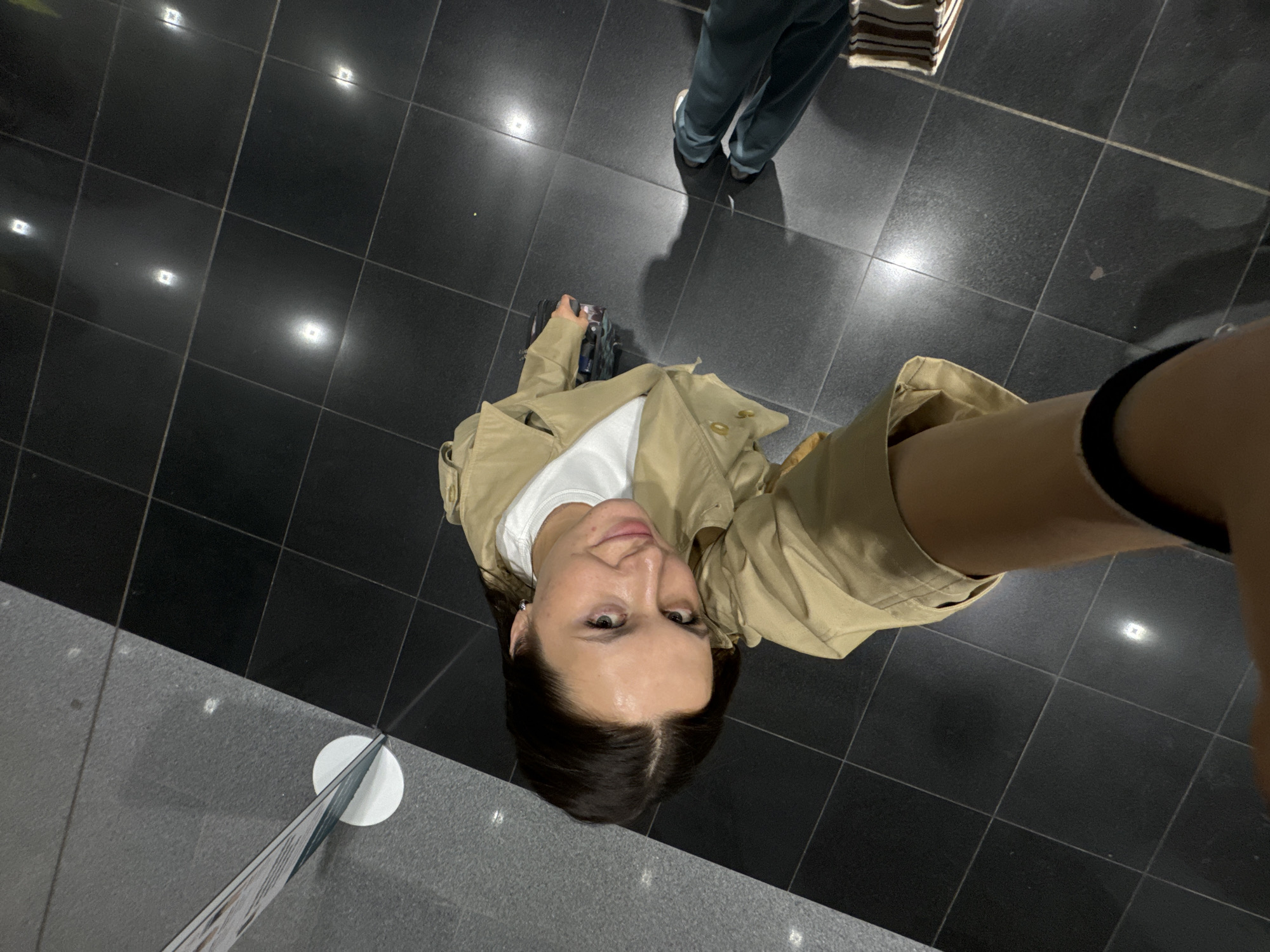 The image is a low-angle selfie taken from a high position, looking down at the photographer. The location appears to be an indoor space with a black tiled floor reflecting light sources above, suggesting a modern, possibly commercial environment. In the bottom left corner, there is a gray tile section with a promotional sign.
The photographer is a fair-skinned woman with dark hair, wearing a light brown trench coat over a white shirt. She is holding a black bag or suitcase in her left hand. In the upper part of the image, partially visible is another person's legs, wearing light green pants and white shoes.