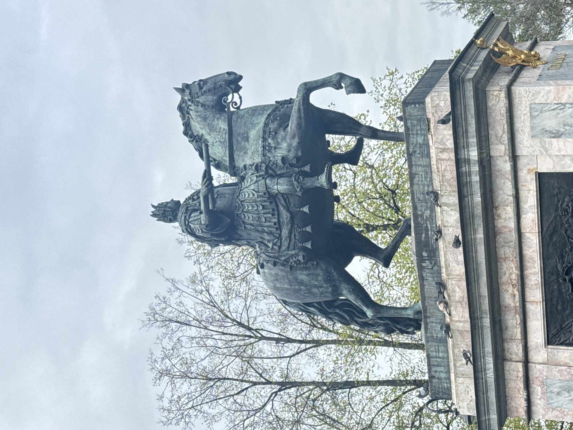 The image shows a low-angle view of the equestrian statue of Nicholas I in Saint Petersburg, Russia. The statue depicts Tsar Nicholas I riding a rearing horse, atop an ornate pedestal composed of marble and bronze. The statue and pedestal are patinated green. Several pigeons are perched on the pedestal. Bare tree branches and budding leaves fill the background, partially obscuring a cloudy sky.