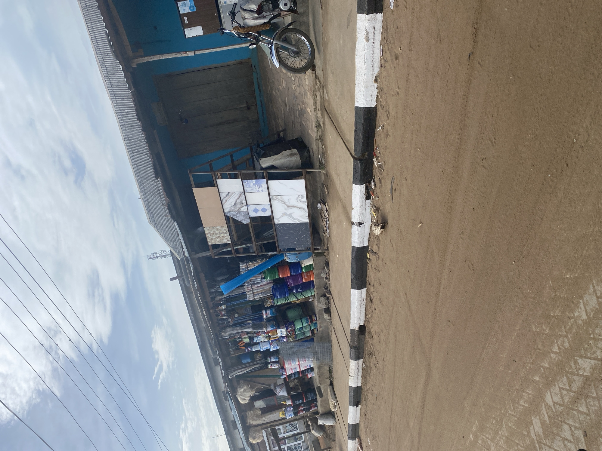 A ground-level, medium shot depicts a street scene in an unidentified urban area. A roadside business establishment occupies the left side of the frame, with a display of construction materials, including tiles and bundles of unidentified items, arranged along the sidewalk. The storefront has a blue facade and a corrugated metal roof. A motorcycle is parked in front of the building. To the right, a dirt road is delineated by a curb painted with alternating black and white stripes. The sky, partially visible in the upper portion of the image, is overcast with light clouds and intersected by power lines.