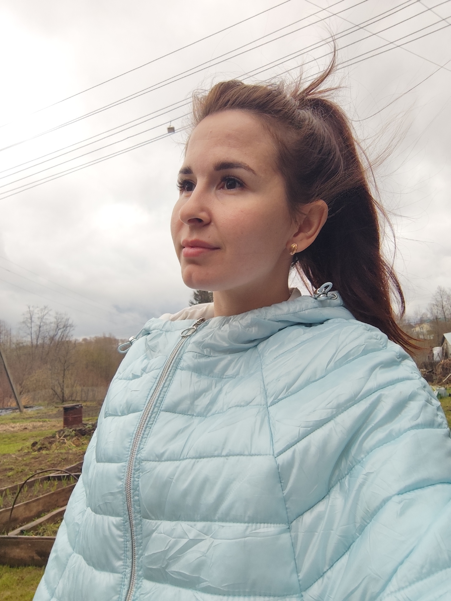 A young woman is pictured outdoors, in what appears to be a rural or suburban area. She is wearing a light blue puffer jacket with a white hoodie underneath and gold stud earrings. Her brown hair is styled in a ponytail, with some strands blowing in the wind.
The background shows an overcast sky with power lines running across it. There is greenery and vegetation, including what seems to be a garden or cultivated area with raised beds. The setting evokes a countryside or a location on the outskirts of a city. The specific city is Фоки, Russia.
