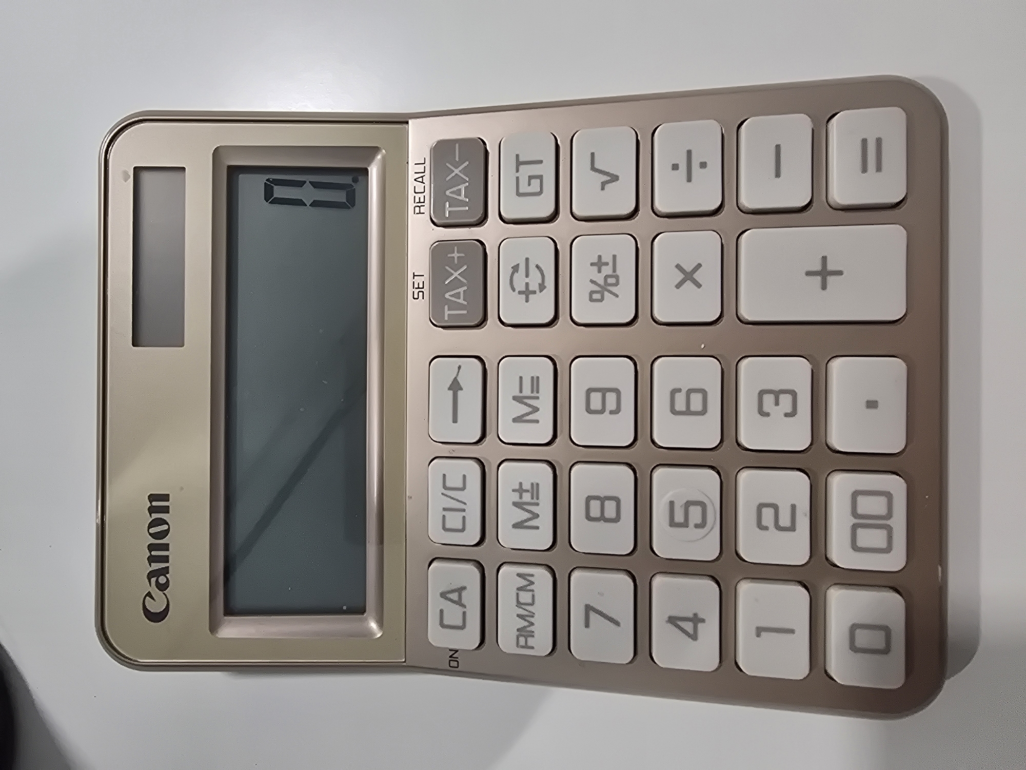The image shows a Canon brand calculator. The calculator is a light brown color with white buttons. The display screen is showing a zero. The brand name "Canon" is printed on the left side of the calculator. The calculator has a standard layout of number and function keys including tax, memory, and percentage functions. There is no indication of a location in Plovdiv, Bulgaria.