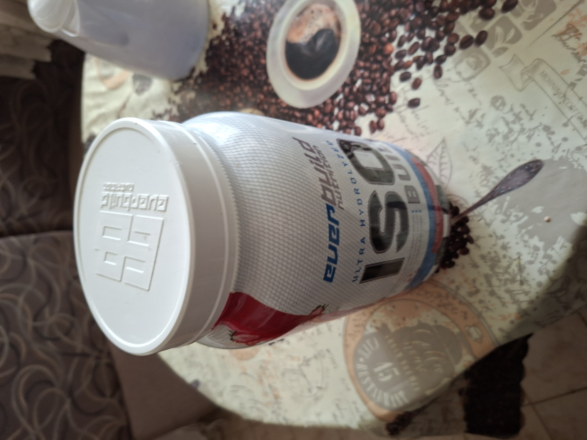 The image shows a container of Everbuild Nutrition Ultra Hydrolyzed ISO Build protein powder on a table with a coffee bean design. A spoon is also visible near the container. Another white plastic container sits in the background. The location is unspecified in terms of Gabrovo, Bulgaria.