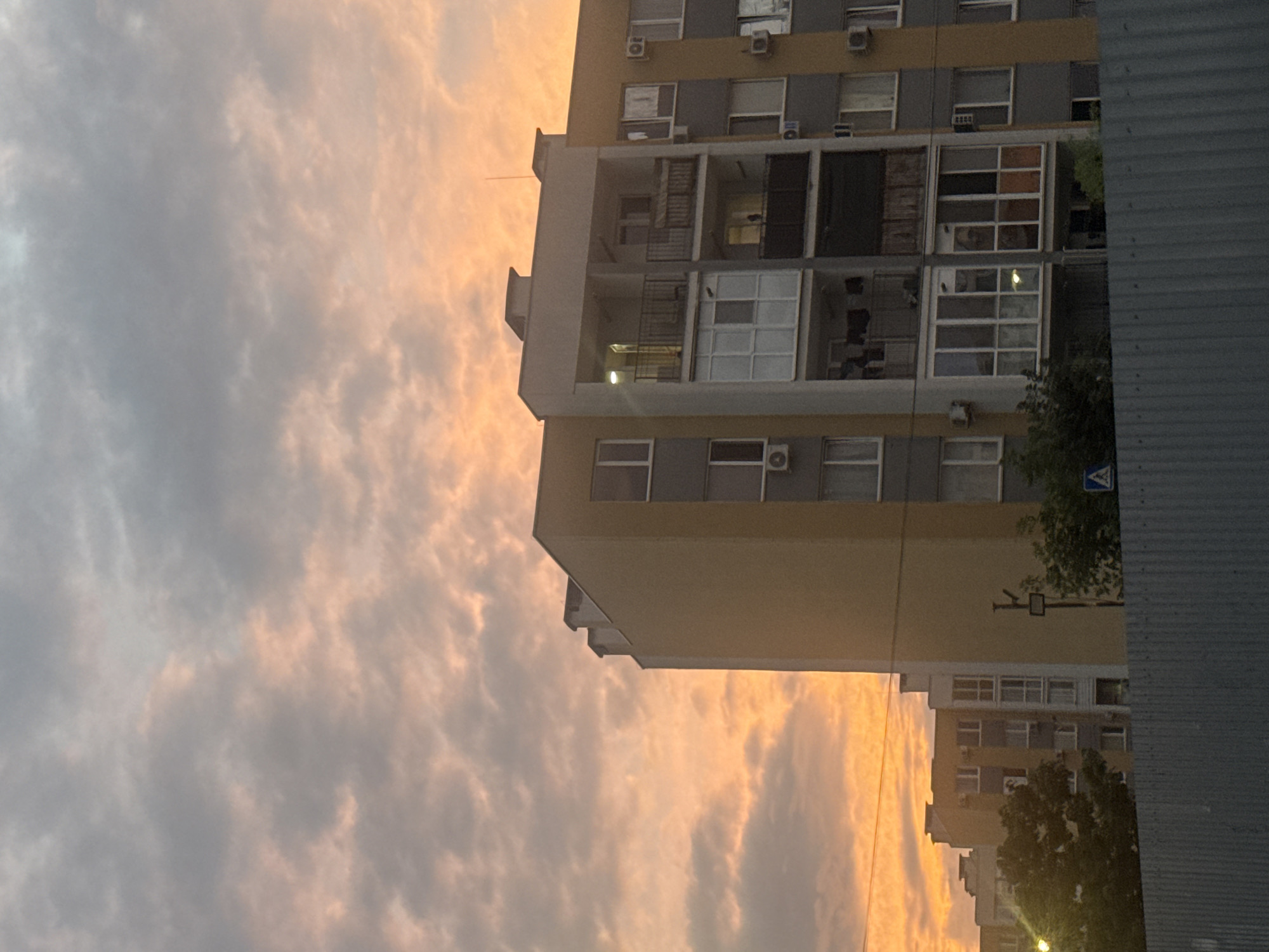 In Avanhard, Ukraine, a building is captured against a dramatic sunset sky. The sky is filled with vibrant orange and pink clouds, with darker grey clouds interspersed. The building, likely an apartment complex, features a yellow facade with grey accents and numerous windows. Balconies are visible on several floors, some with railings and enclosed glass areas. Air conditioning units are mounted on the exterior walls. In the foreground, a grey metal fence runs horizontally across the bottom of the frame, partially obscuring the view of the building and some trees. The lighting suggests it is either sunrise or sunset, casting a warm glow over the scene.