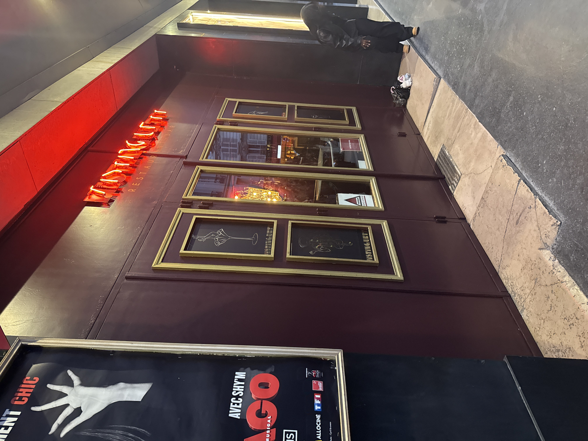 This oblique, eye-level shot captures the illuminated facade of a building, likely a restaurant or bar, on an urban street in Paris, France, during what appears to be dusk or evening. The dominant feature is a vibrant red neon sign displaying "MISTINGUETT" in large letters, with "RESTAURANT BAR" in smaller vertical text below, set against a dark red/maroon exterior.
The building's facade features several gold-framed elements. At the top right, two smaller frames contain stylized illustrations, with "MISTINGUETT" also visible beneath them. Below these are two larger, rectangular windows, reflecting street elements and containing a small "FUMEURS INTERDITS" (Smokers Forbidden) sign.
In the bottom left foreground, a large, silver-framed poster is partially visible. It advertises a musical, with text including "NT CHIC", "AVEC SHY'M", and "GO MUSICAL". Logos for "ALLOCINE", "TF1", and "inter" are also present, along with a credit for "Photo: Cyril Bruneau". A prominent graphic of a white, disembodied hand is centered on the poster.
On the right side of the image, a person dressed in dark clothing and tan shoes stands on the sidewalk, their back mostly to the camera, next to a small white bag or item. The sidewalk consists of light-colored cracked tiles and darker asphalt, with a metal grate visible in the foreground. No specific activity is clearly taking place beyond the person standing.