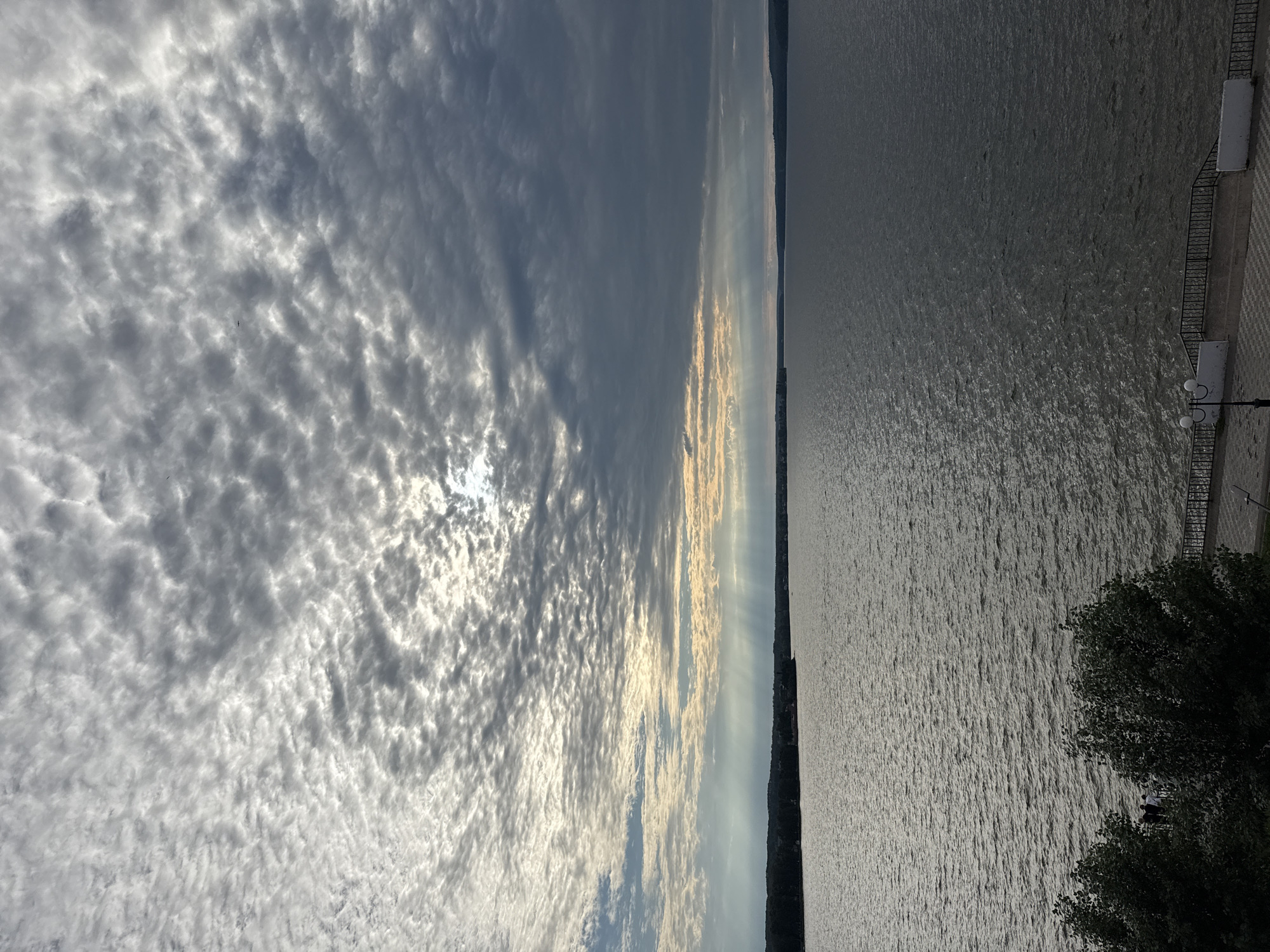 The image shows a vast body of water, likely the Votkinskoye Reservoir, on a cloudy day. Sunlight streams through gaps in the clouds, creating dramatic light shafts that illuminate the water and the distant shoreline. The shoreline is lined with trees and appears to be a city or town. In the foreground, there are trees and a paved walkway with a railing, suggesting a public area like a park or embankment. A lamppost is visible on the walkway. The overall impression is a serene and somewhat moody landscape, characteristic of a northern climate. The location is Votkinsk, Russia.