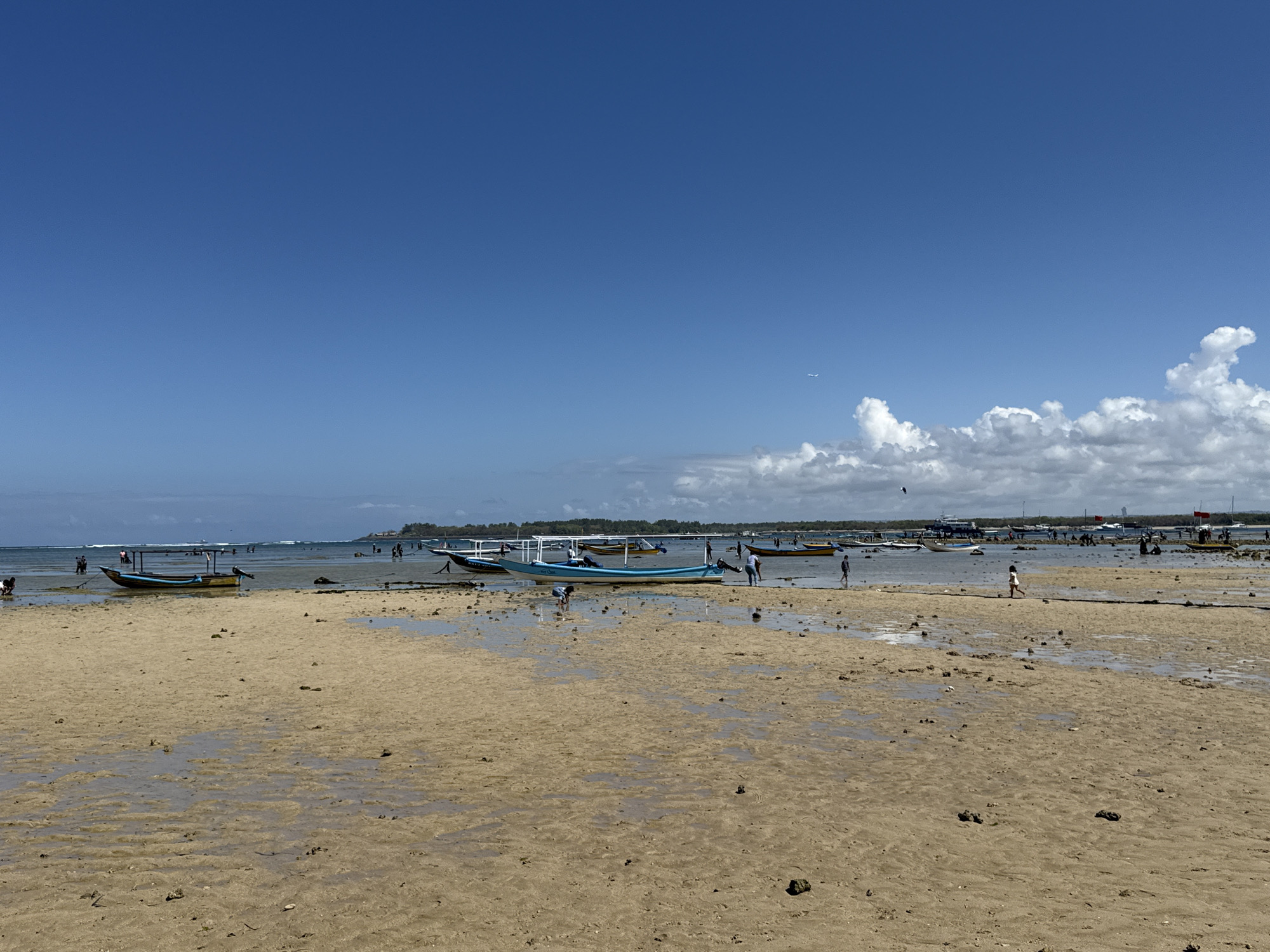 This is a daytime, eye-level, wide-angle shot of a beach scene at low tide in Denpasar, Indonesia. The foreground is dominated by wet sand with small puddles and scattered debris. In the middle ground, several traditional Balinese jukung boats are beached or partially submerged in shallow water. Some boats are blue and white, others yellow and blue, and they are of varying sizes. A few people are visible walking on the sand, near the boats, and in the shallow water, some appearing to be interacting with the boats. In the background, the ocean stretches to the horizon under a clear blue sky with scattered white clouds. More boats and a distant shoreline with trees are visible. An airplane is visible in the sky above the clouds. The overall scene depicts a typical beach environment with boats and people engaged in various activities.