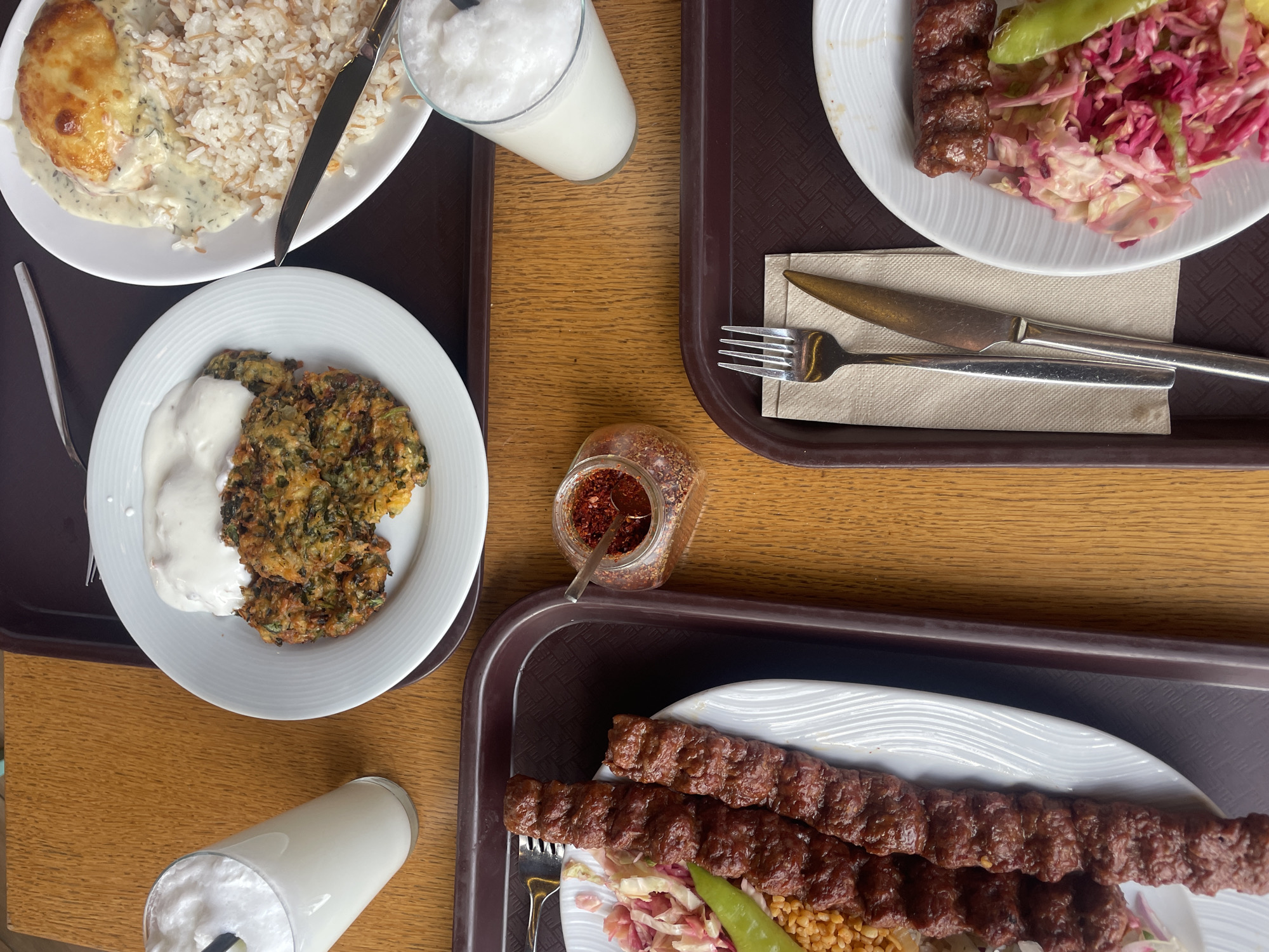 An overhead shot displays two meals arranged on brown plastic trays atop a light brown wooden table in a casual dining setting. The precise location within Sofia, Bulgaria, is not identifiable from the image.
The upper tray contains a white plate with three round, pan-fried green vegetable fritters, topped with a dollop of white creamy sauce. Adjacent is another white plate holding a portion of rice pilaf with vermicelli, beside a baked dish featuring a protein (likely poultry or meat) covered in a creamy herb sauce and melted, browned cheese. A silver knife rests diagonally on the latter plate. Two tall glasses, one partially obscured, contain a foamy white beverage, possibly ayran; one includes a black straw.
The lower tray prominently features a white plate with two skewers of dark brown, ground meat kebab. This is accompanied by a portion of light brown bulgur pilaf, a shredded red and white cabbage salad, and a single whole green pepper. A silver fork rests on the upper left edge of the kebab plate. A small, clear glass jar with a metal spoon, containing red pepper flakes, is situated between the two trays. In the lower right corner of the image, on the lower tray, a set of silver fork and knife rests on a beige napkin. No individuals are depicted.